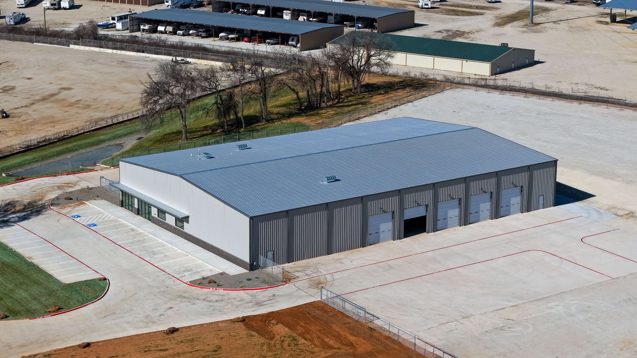 Aerial view of a large industrial warehouse with a metal roof and multiple garage doors, surrounded by concrete pavement and parking spaces.