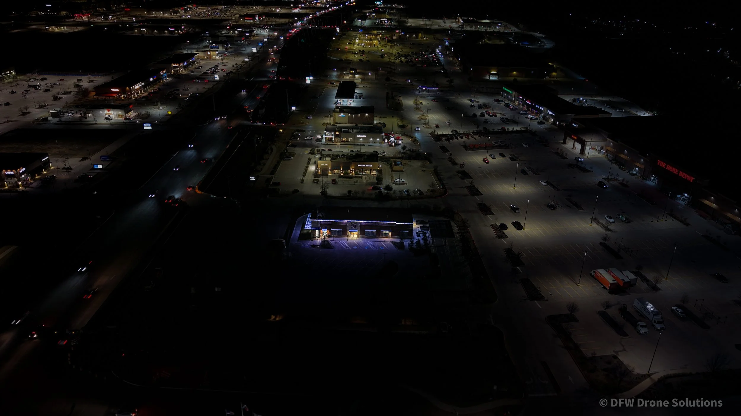 Aerial night view of a shopping center with well-lit parking lots and a visible Home Depot store. Several other commercial buildings are illuminated, and streets with moving vehicles can be seen.