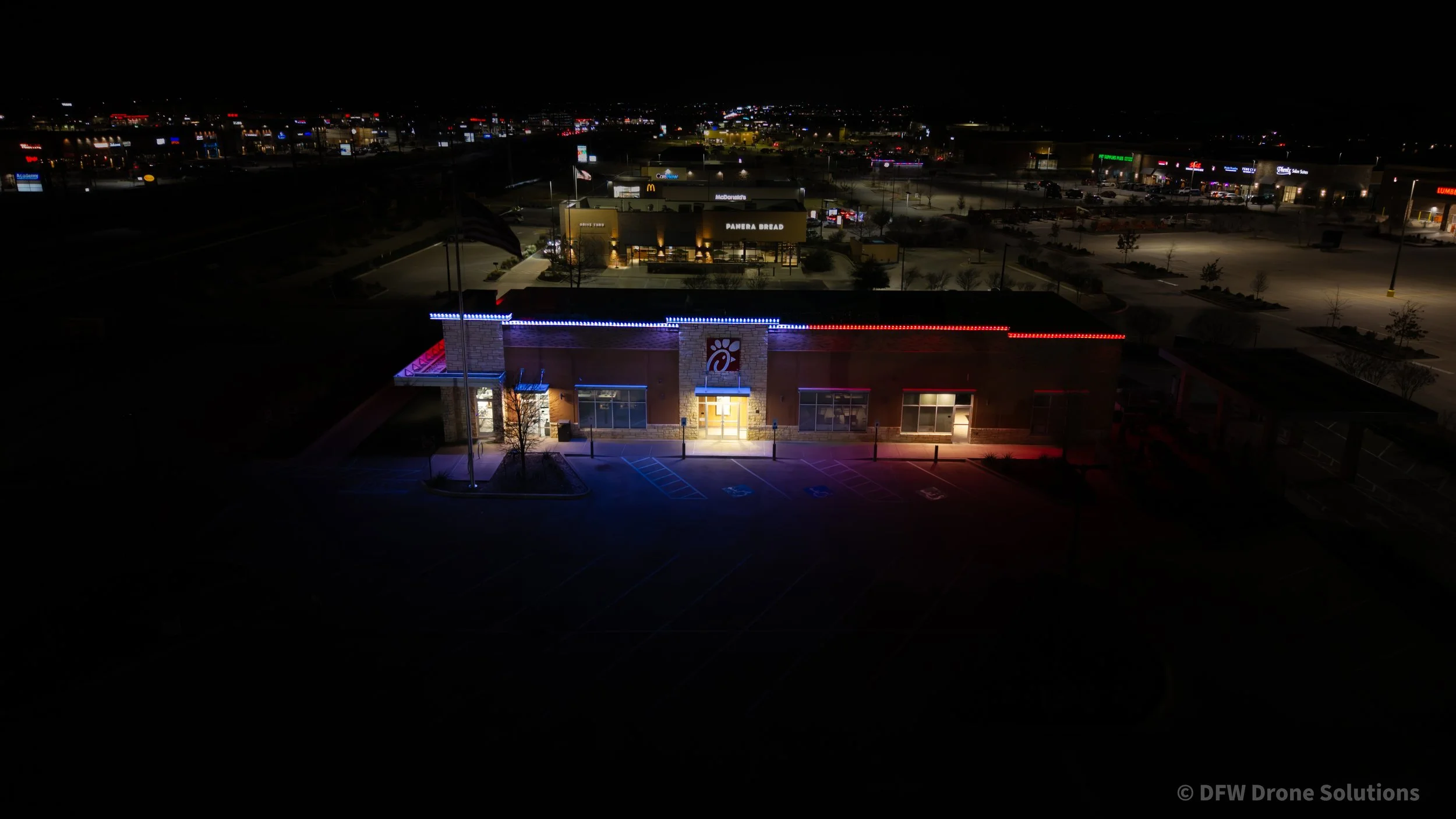 Night aerial view of a Chick-fil-A restaurant with illuminated sign and red-blue lights, located in a commercial area with visible neighboring stores and parking lot.