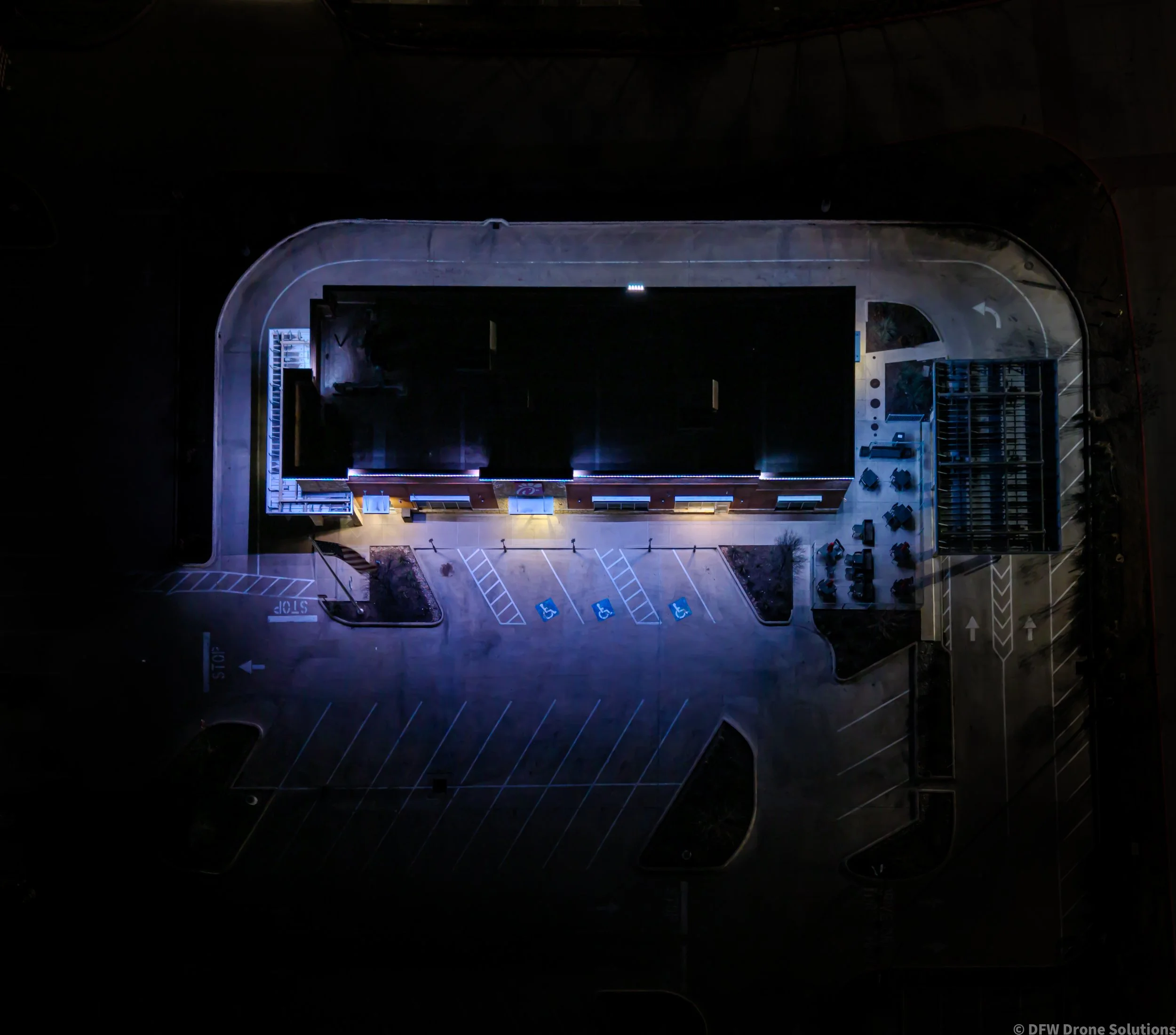 Aerial night view of an illuminated building with surrounding parking lot, featuring marked parking spaces, including handicap spots, and visible pedestrian pathways.