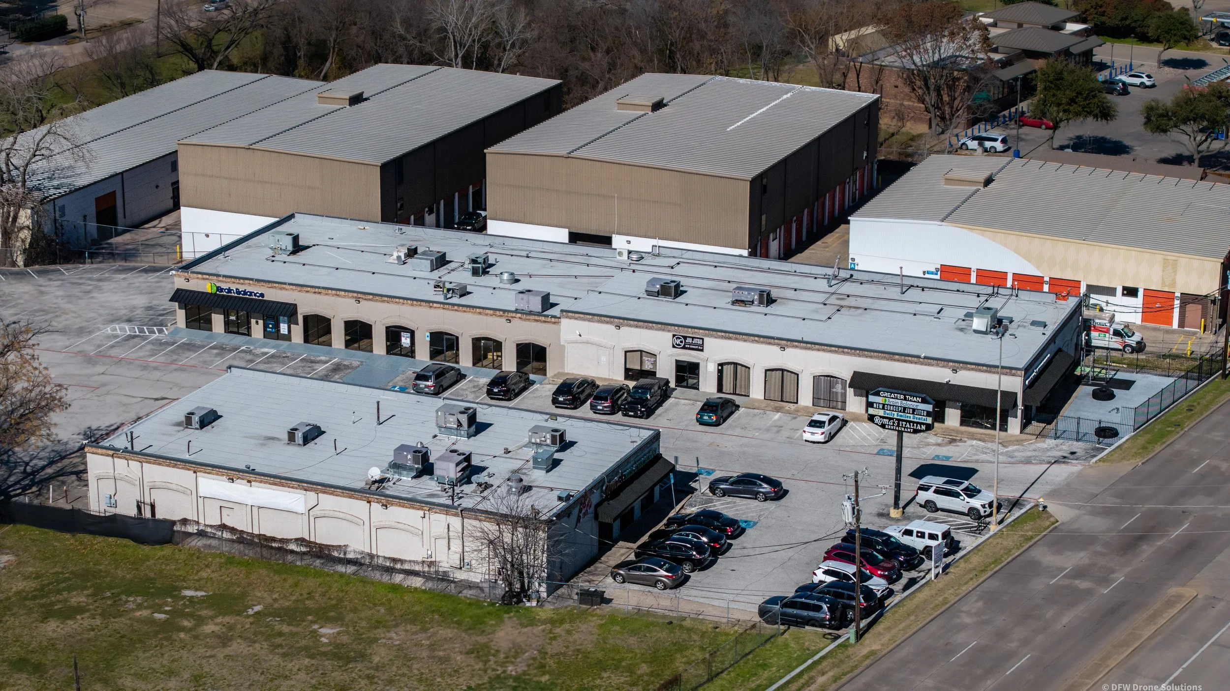 Aerial view of a commercial shopping center with multiple businesses, parking lot with cars, and surrounding buildings.