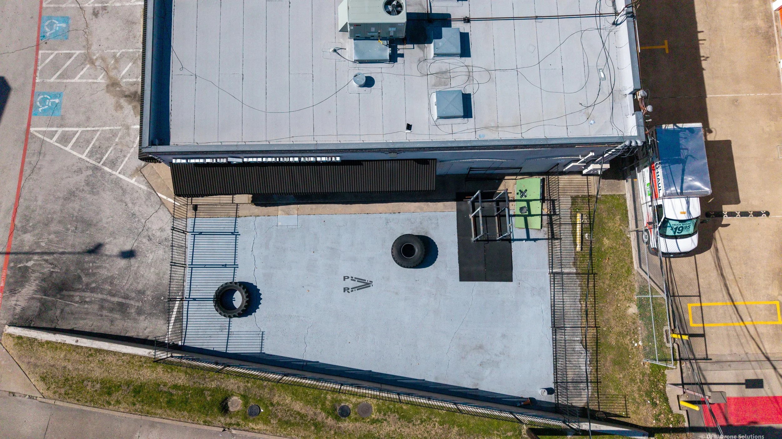 Aerial view of building with parking lot, rooftop HVAC units, and enclosed outdoor workout area with tires and gym equipment.