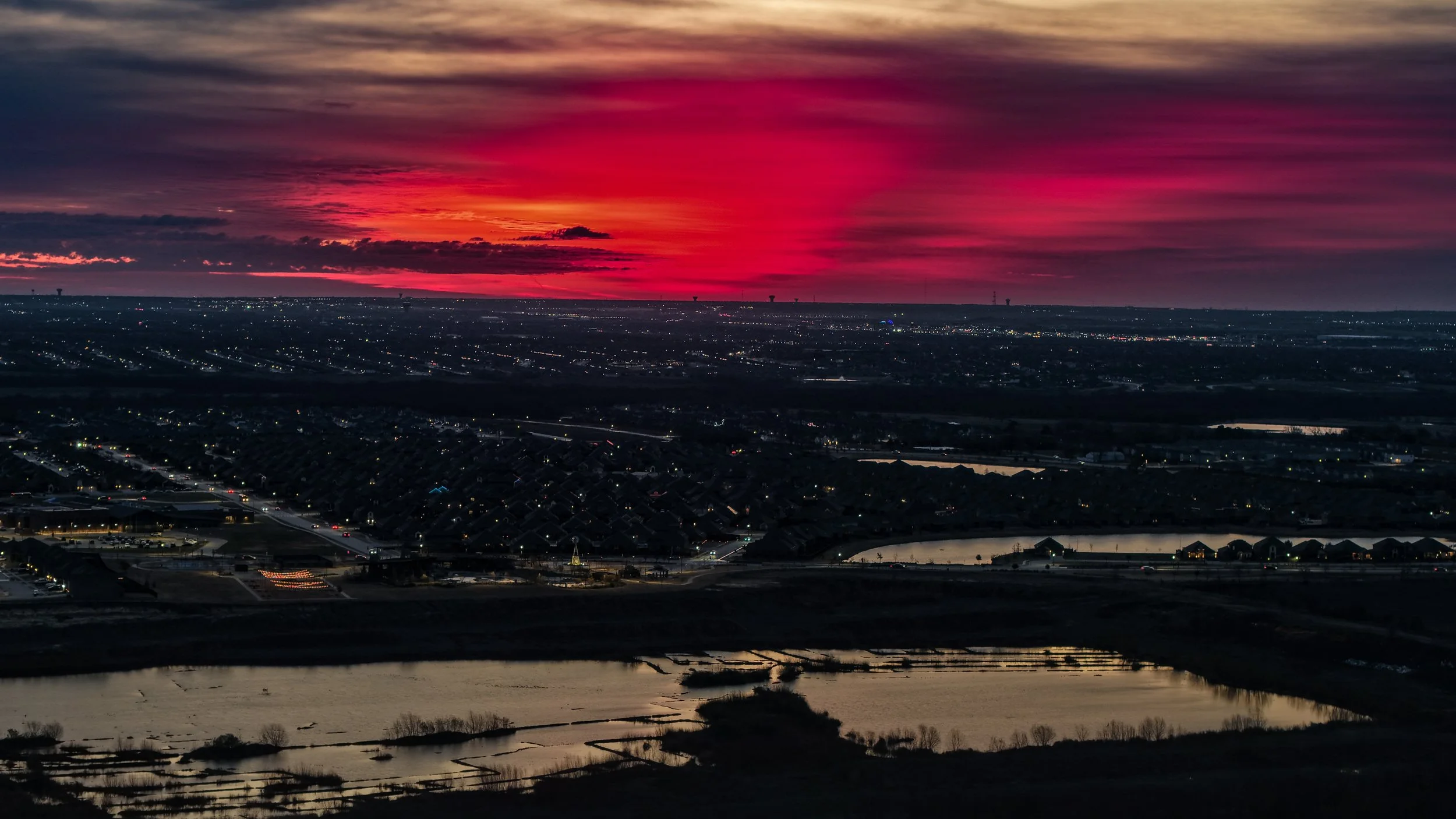 Aerial view of a cityscape at sunset with a vibrant red and pink sky, urban area with lights, residential neighborhood, and water bodies in the foreground.