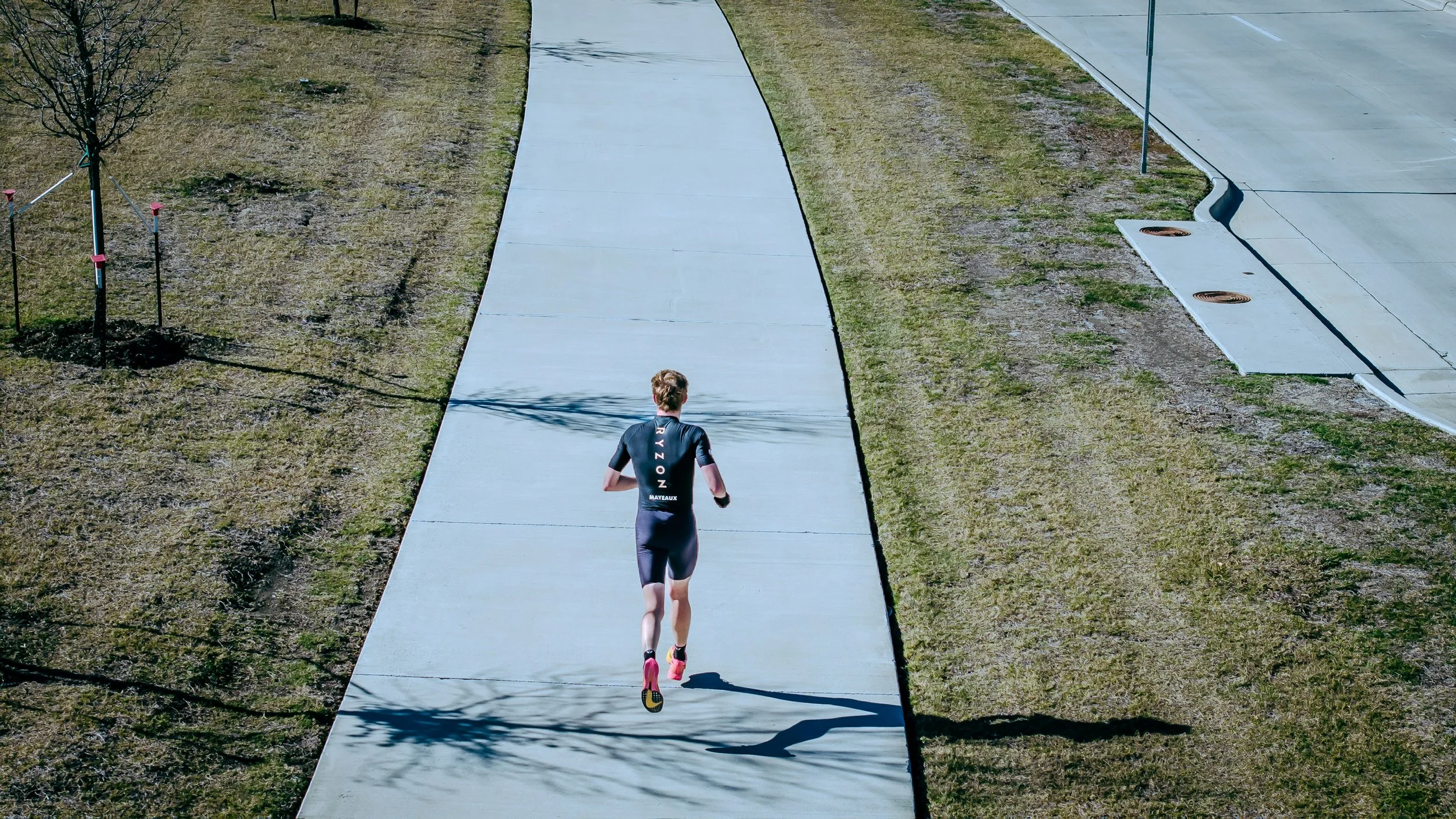 An individual running on a paved path surrounded by grass with trees and a road nearby.