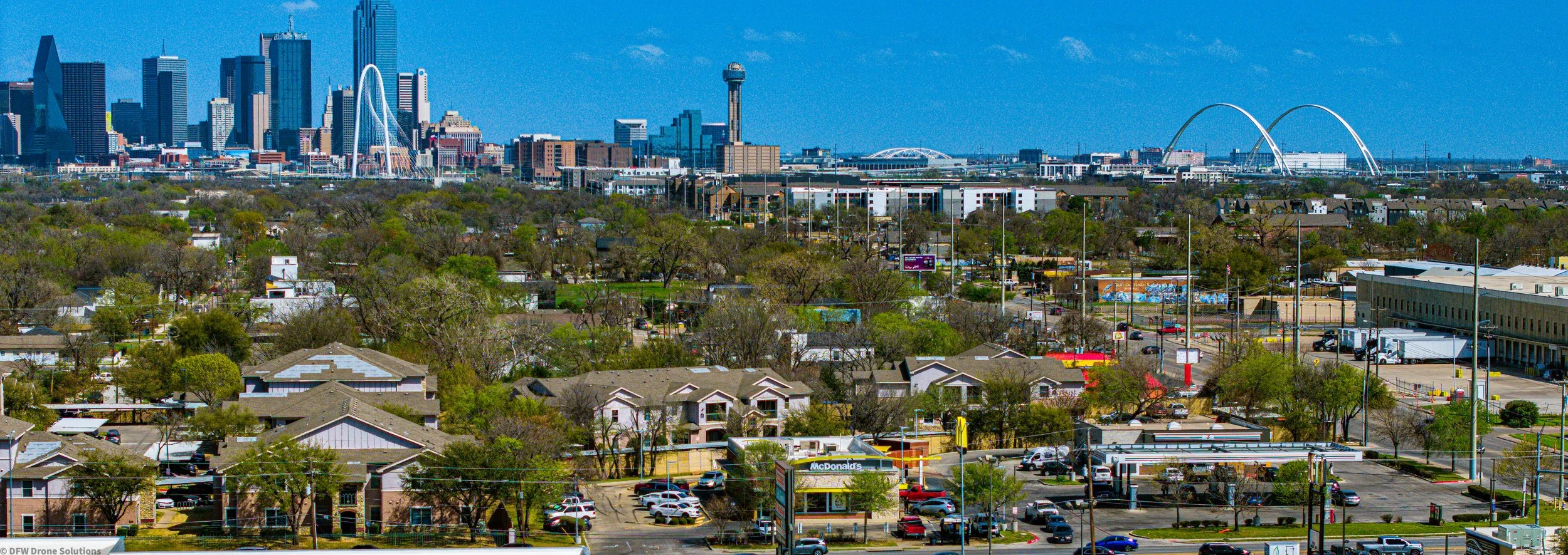 Aerial view of Dallas, Texas skyline with residential areas and greenery in the foreground. Prominent skyscrapers and landmarks like the Reunion Tower and Margaret Hunt Hill Bridge are visible.
