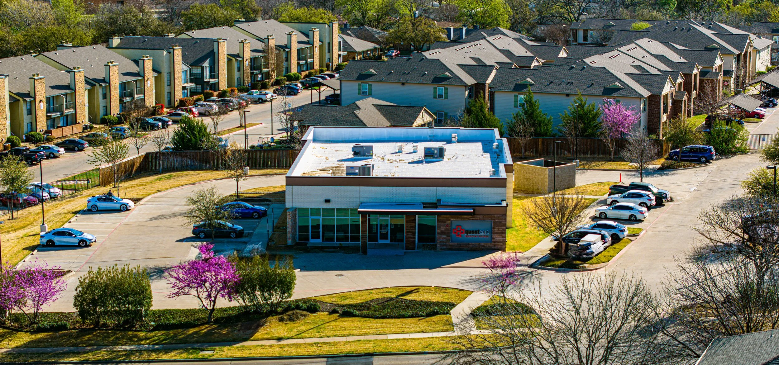 Aerial view of a commercial building with a parking lot, surrounded by residential houses and trees. Cars are parked in the lot, and blooming trees add color to the scene.