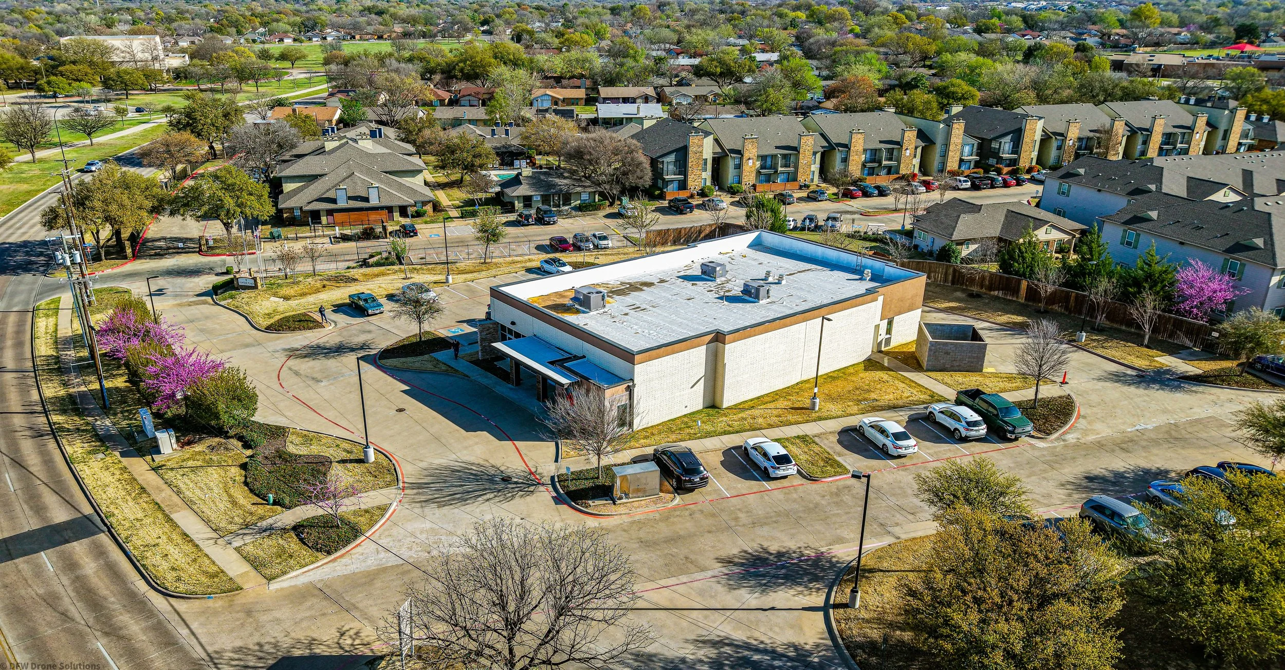 Aerial view of a commercial building with a white roof and surrounding parking lot in a suburban neighborhood. The area has leafy green trees, adjacent residential houses, and a paved roadway. Several cars are parked around the building, and pink flowering trees are visible along the pathways.