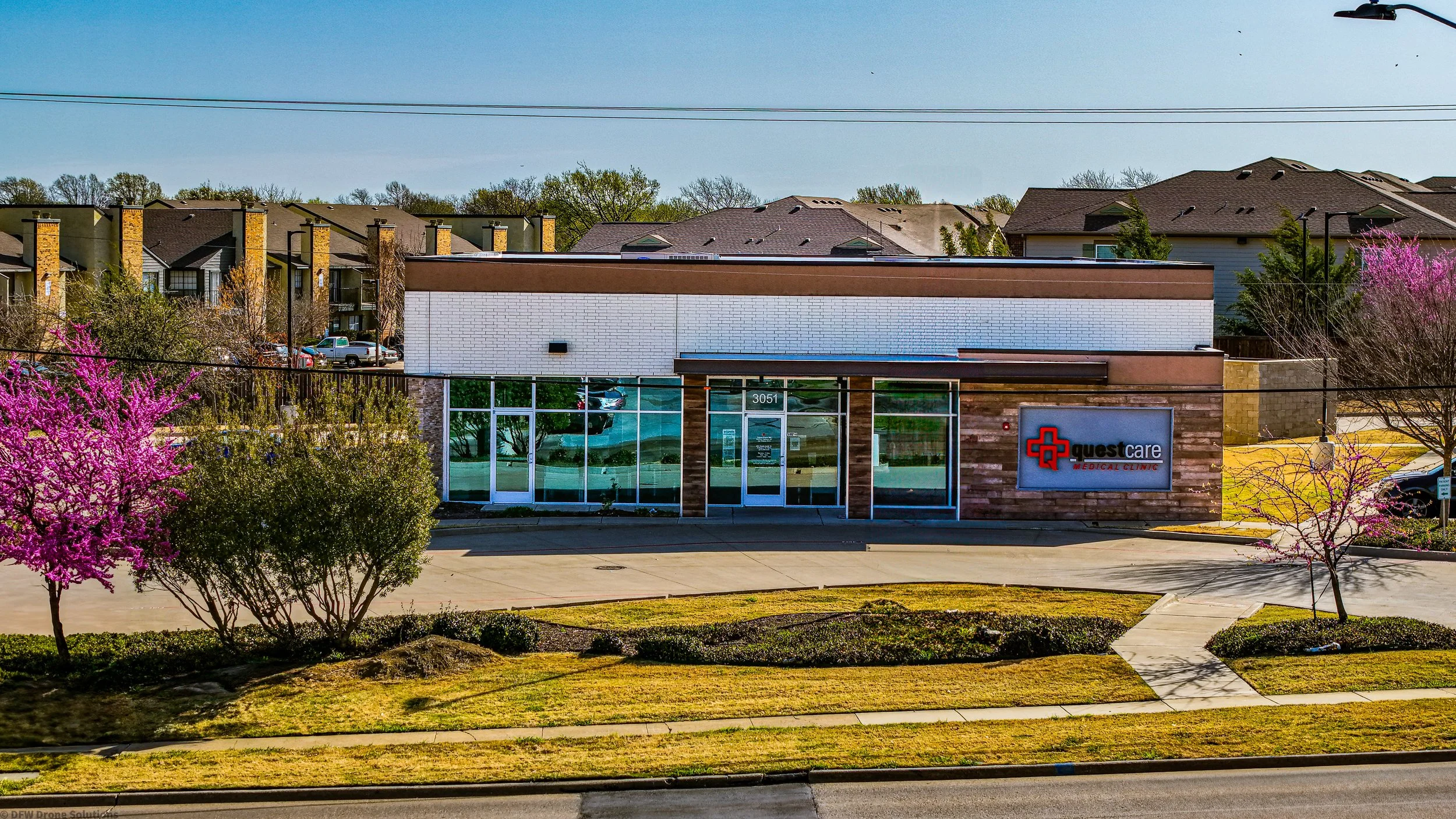 Medical clinic building with a sign reading 'Questcare Medical Clinic,' surrounded by trees and grass, under a clear blue sky.
