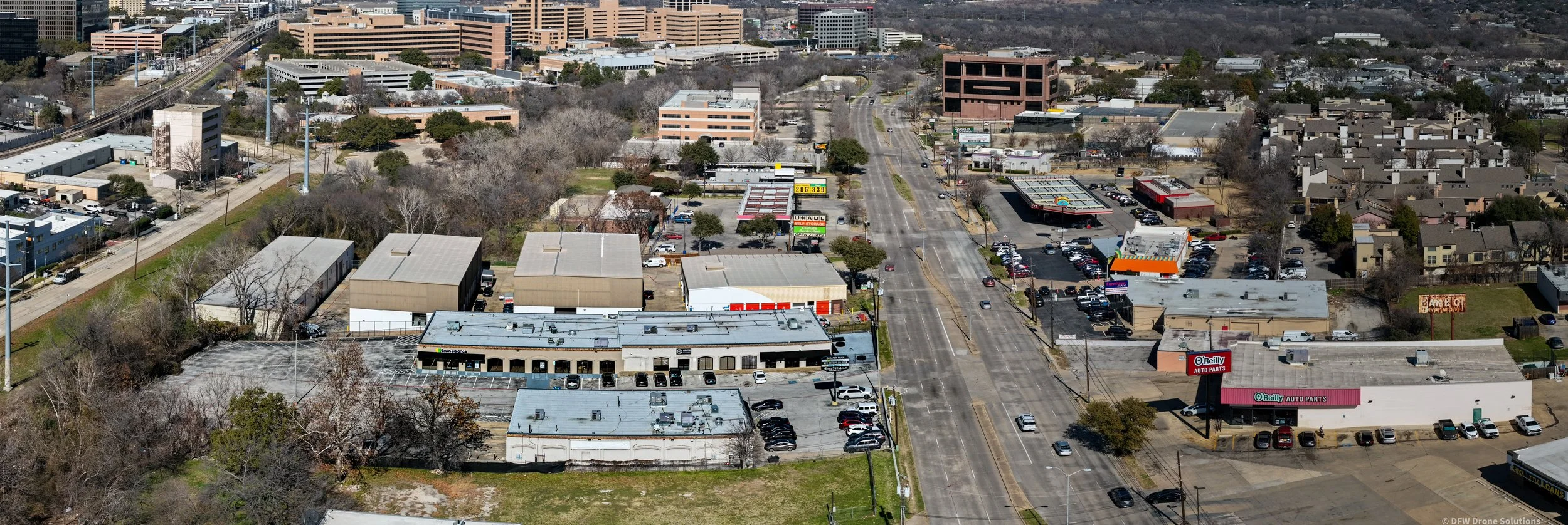 Aerial view of an urban area with a wide street lined with various commercial buildings, parking lots, and a few cars. The landscape includes some trees and an arrangement of larger, institutional structures further in the background, possibly part of a business or medical district. Signs for businesses like auto parts and other stores are visible.