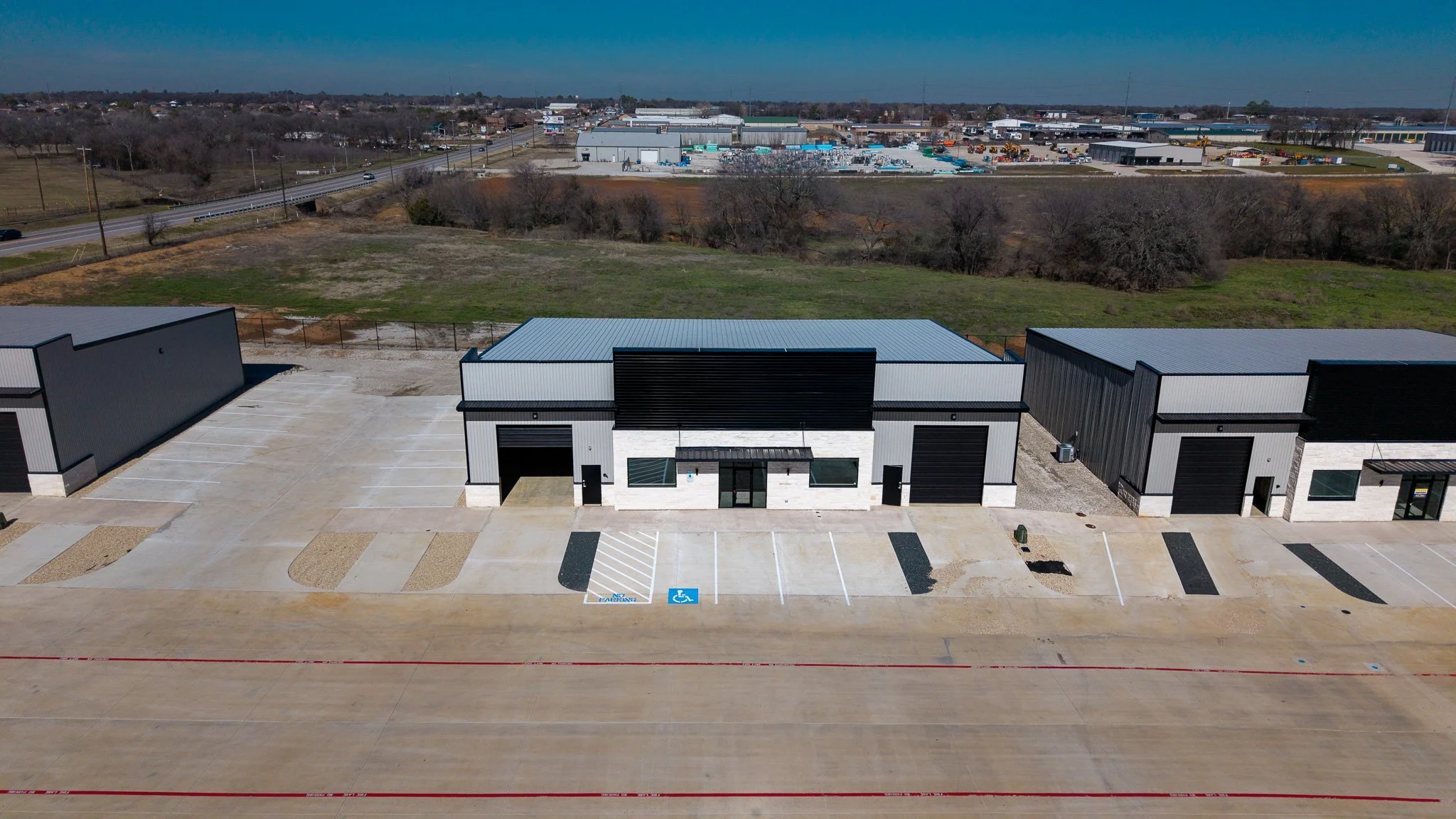 Industrial complex with three gray warehouse buildings, parking lot, and a highway in the background.