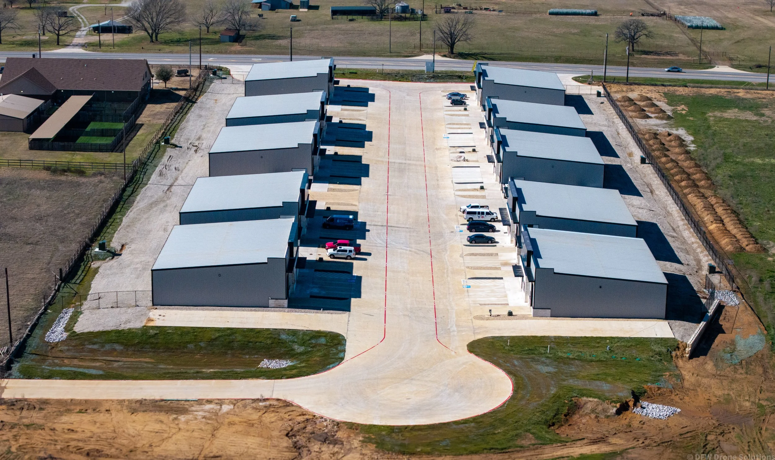 Aerial view of a row of industrial storage units with parking spaces and several parked vehicles in a rural area.