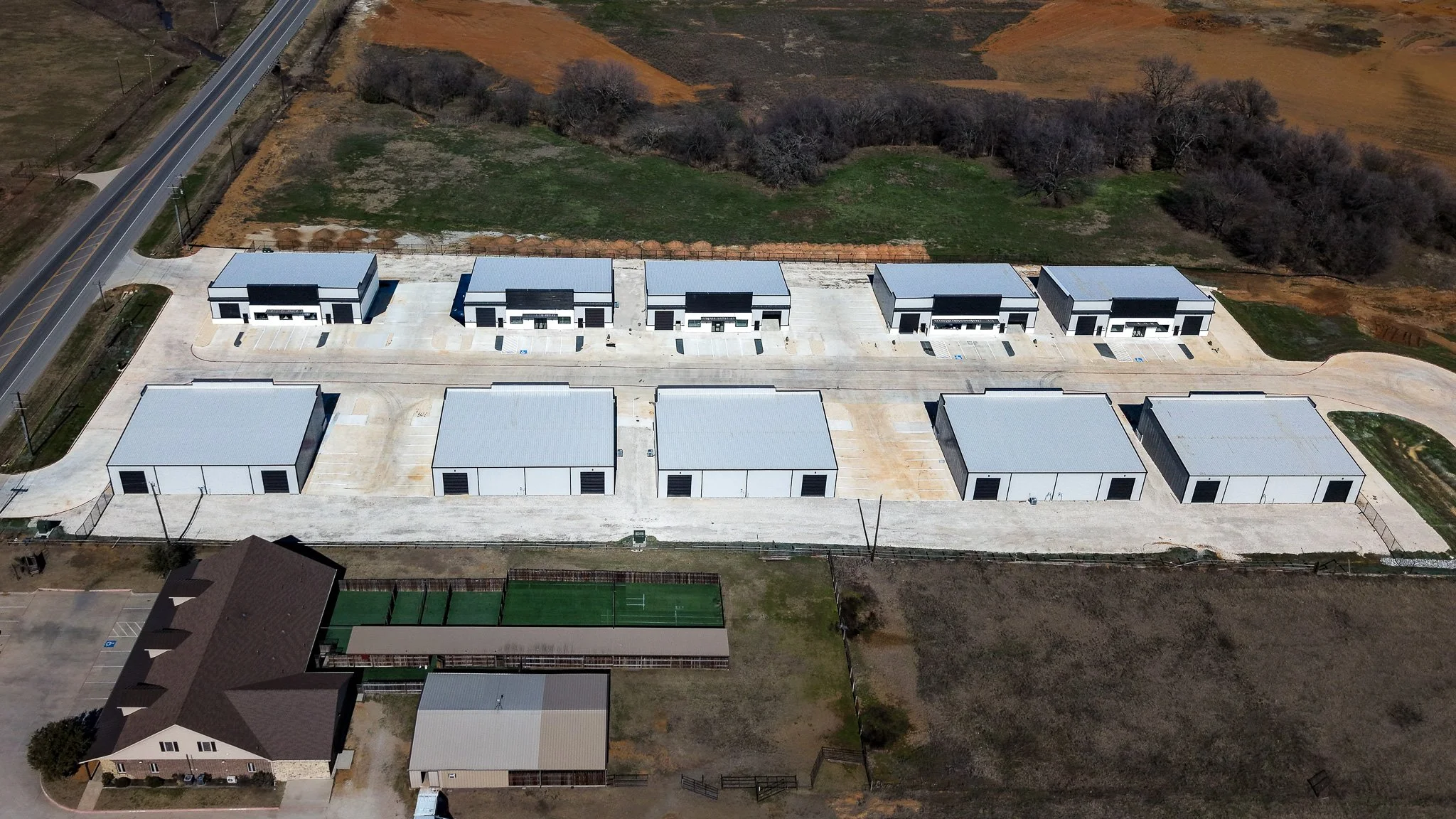 Aerial view of industrial warehouse buildings with parking lots adjacent to a rural road and surrounded by grass and dirt areas.