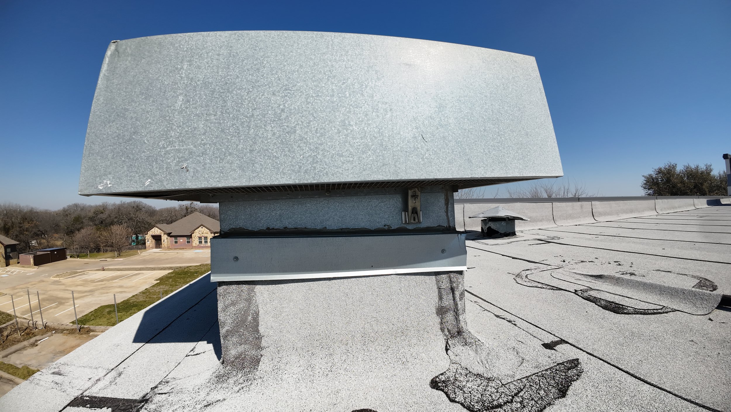 Metal vent on a flat rooftop under a clear blue sky, with nearby buildings visible.