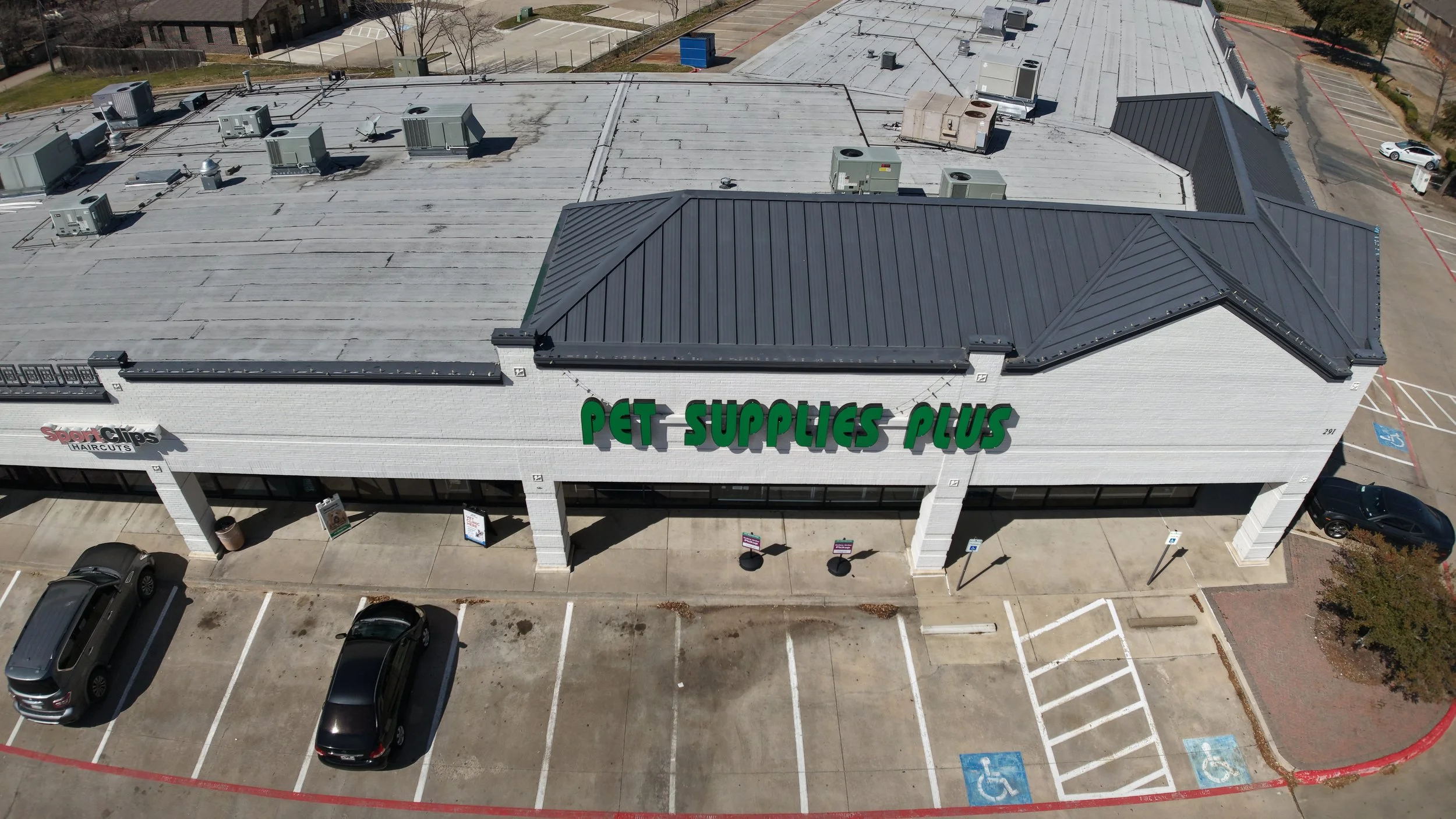 Aerial view of a shopping center with Pet Supplies Plus and Sport Clips stores, visible parking lot with cars.