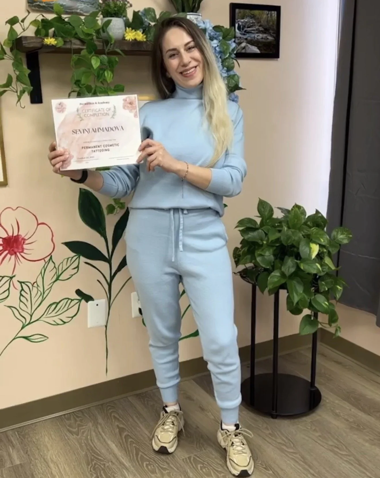Woman holding a certificate of completion for permanent cosmetic tattooing, standing in a room with floral wall art and potted plants.