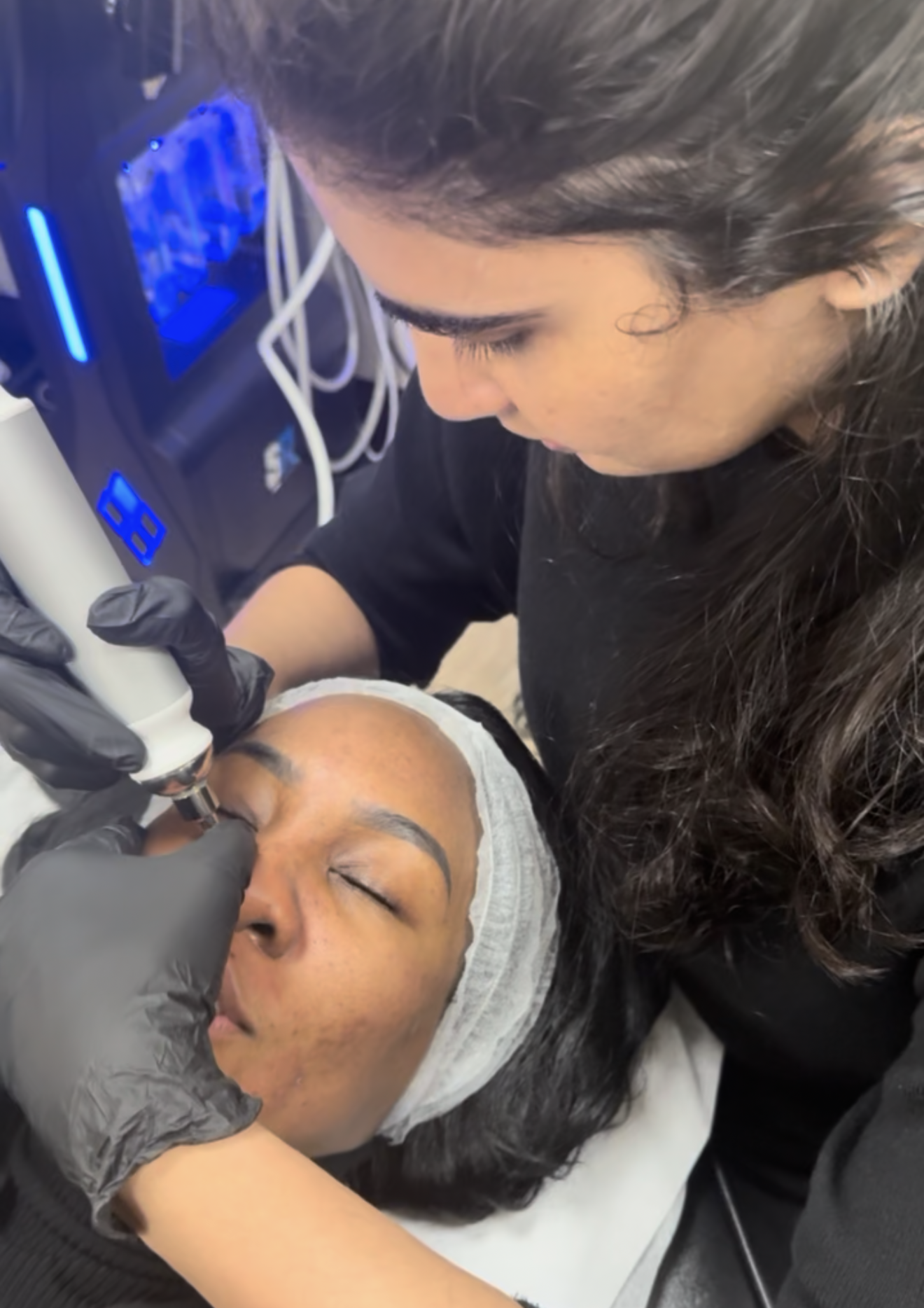 A woman receives a cosmetic procedure on her eyebrows while lying down with a headband and a disposable cap. A technician, wearing black gloves, uses a device on her forehead. The setting appears to be a clinic or spa with electronic equipment and wires in the background.