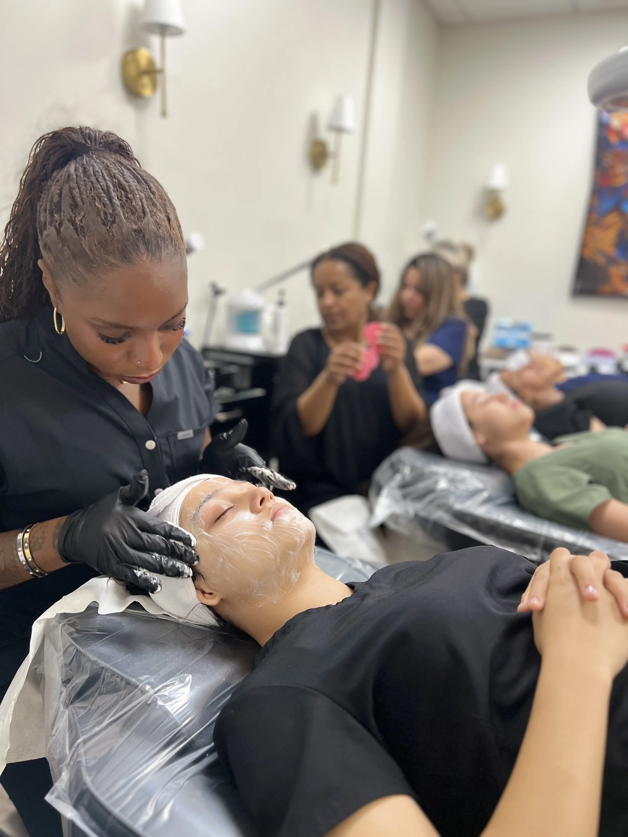 A woman getting a facial treatment at a spa or beauty clinic, lying on a treatment bed with a face mask applied. Other people are in the background, some sitting and one taking a photo.
