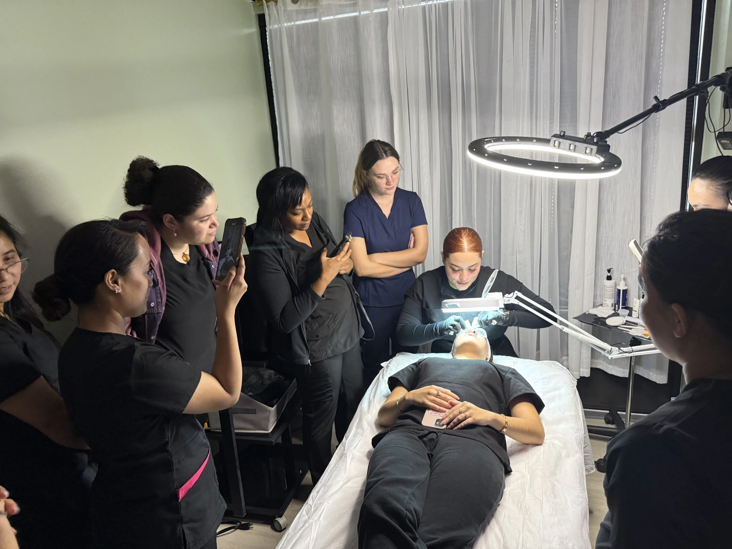 A group of women, some with smartphones, observing a woman performing a procedure on a patient lying on a medical bed. The practitioner uses bright lighting and wears gloves, with medical supplies on a nearby tray.