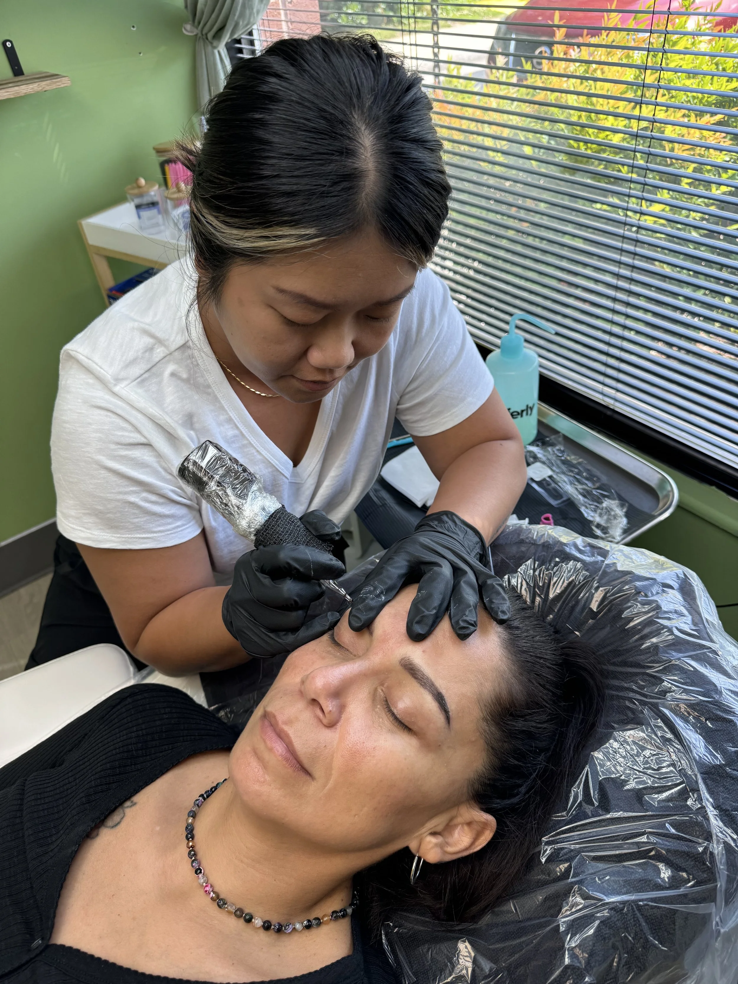 A woman lying down with her eyes closed receives a cosmetic tattooing procedure on her eyebrow from a professional artist wearing black gloves in a skincare clinic.