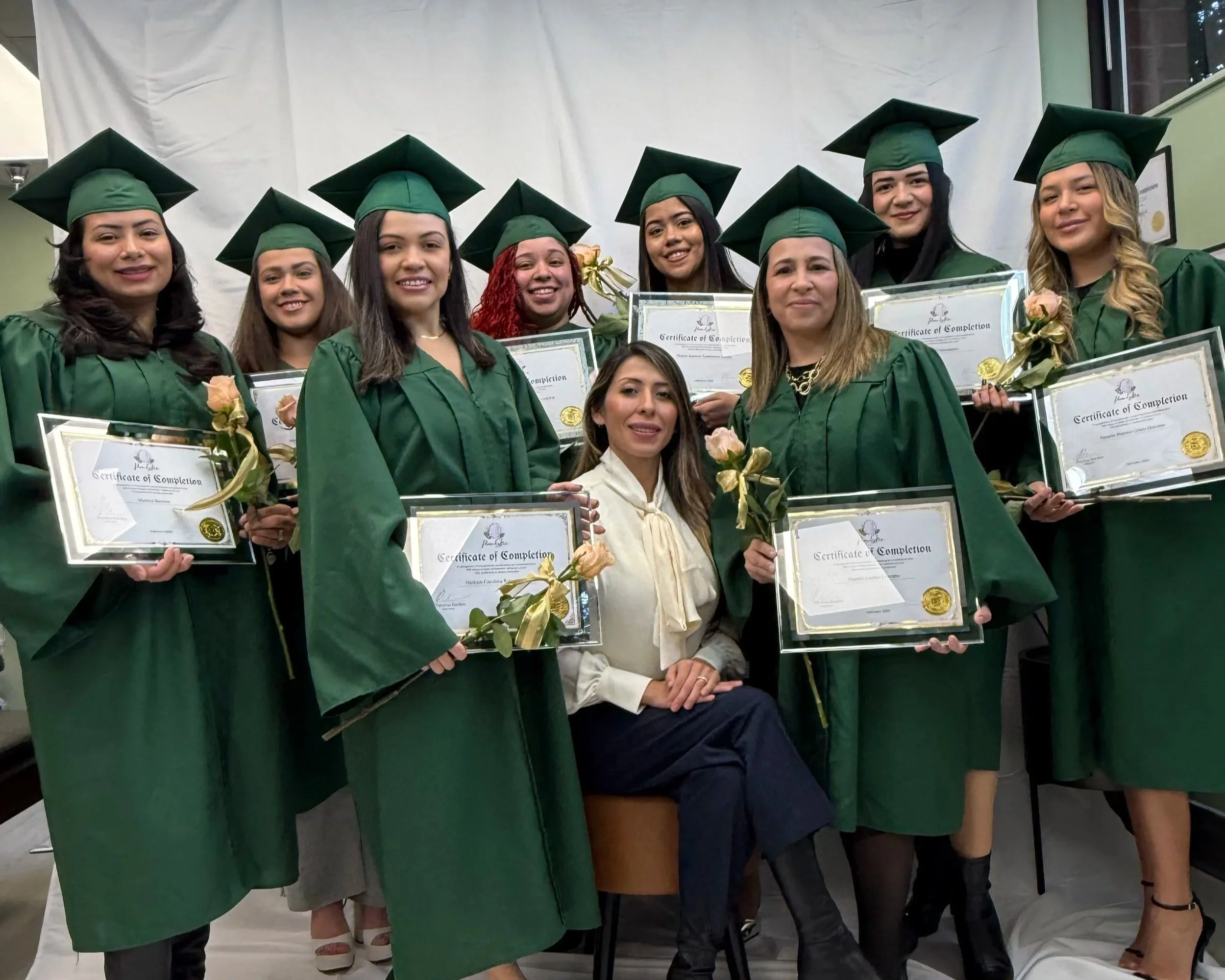 Group of women in green graduation gowns and caps holding diplomas and flowers, posing with a seated woman in beige attire, against a white background.