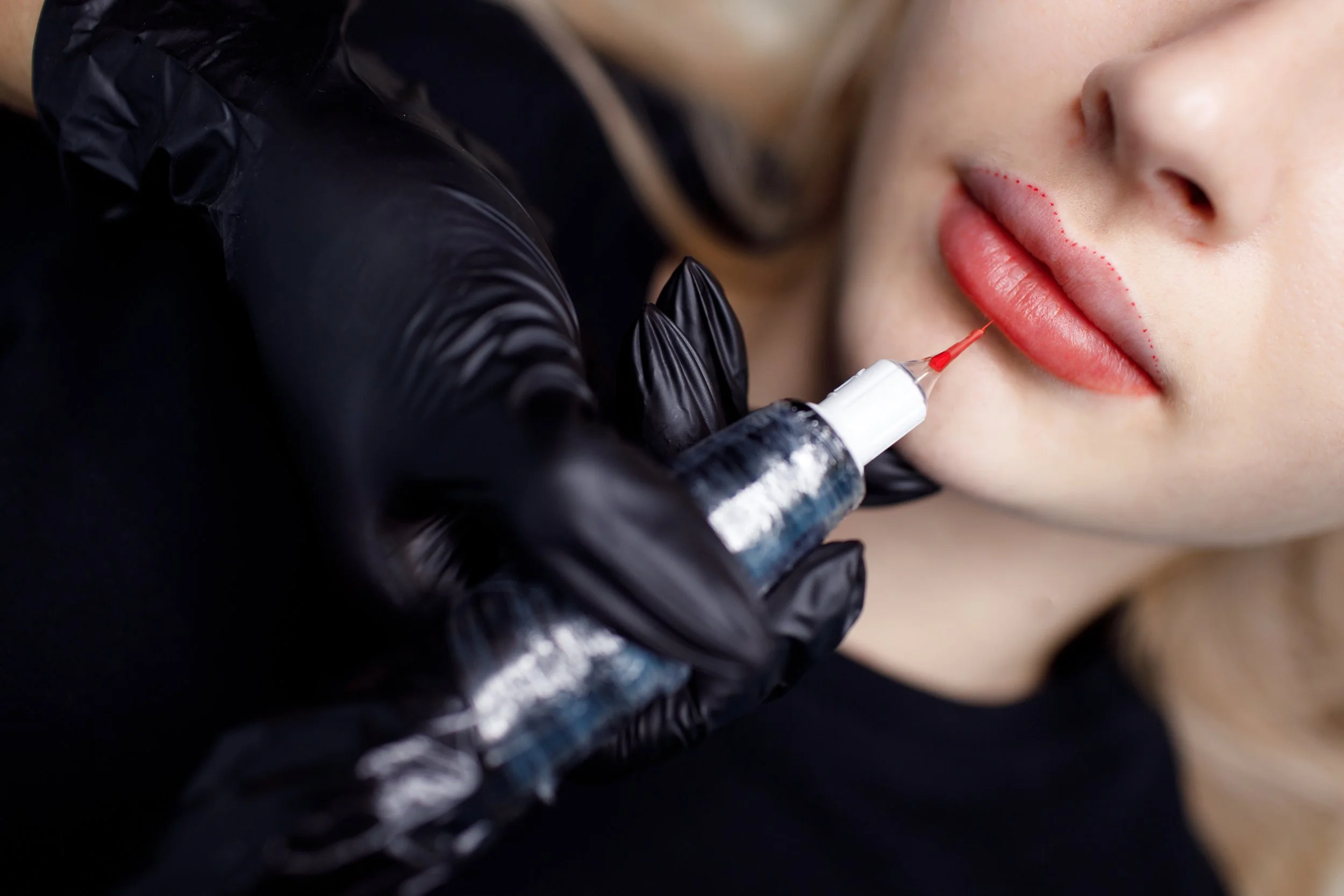 A person receiving a lip tattoo with a tattoo syringe, wearing black gloves, with dotted marks outlining the lips for the procedure.