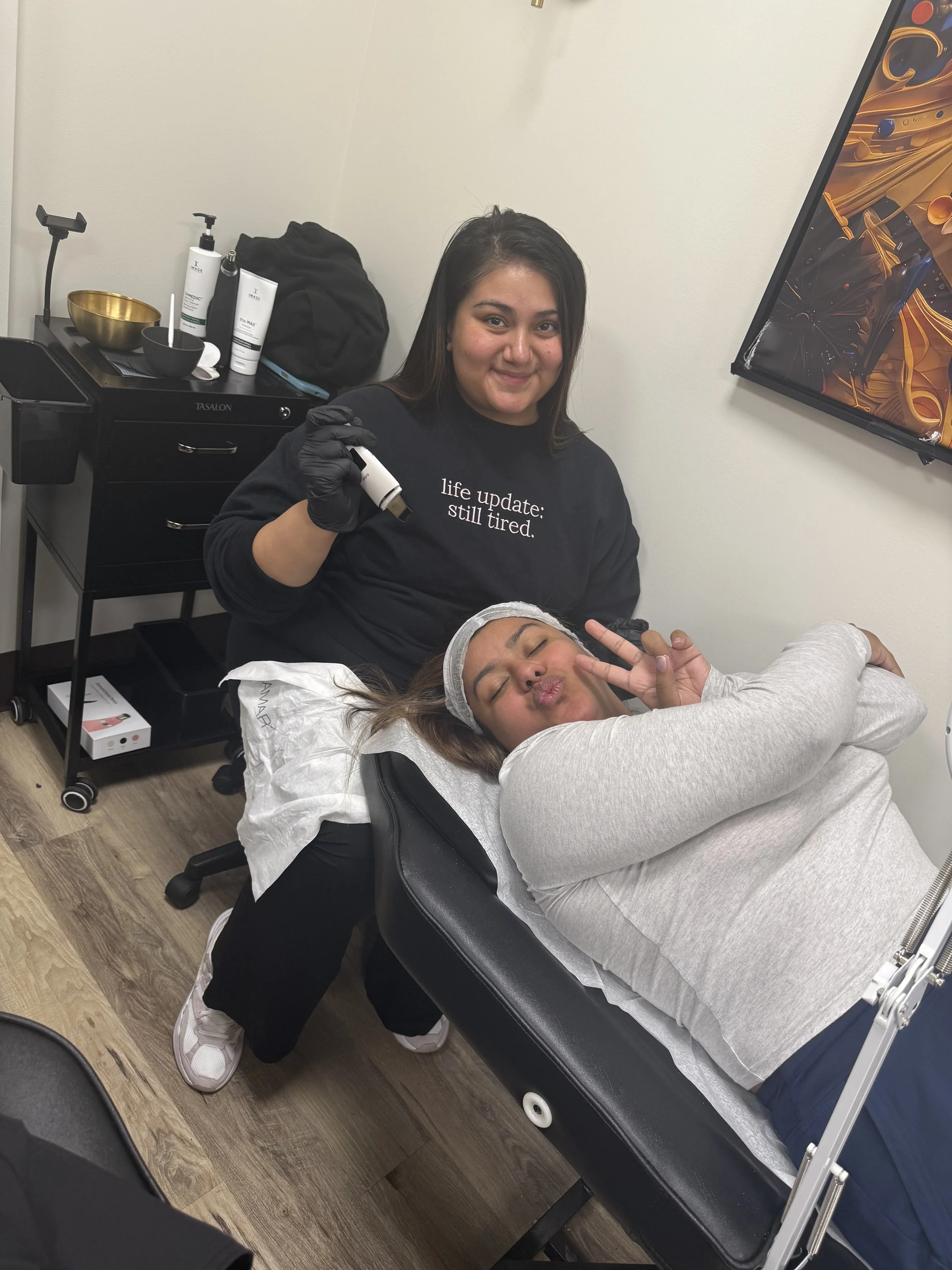 A woman lying on a treatment bed making a peace sign with her fingers and puckering her lips, while another woman standing next to her holds a small device in her hand and smiles. The woman lying down is wearing a hair cap and gloves, indicating she is receiving a cosmetic or skincare treatment.