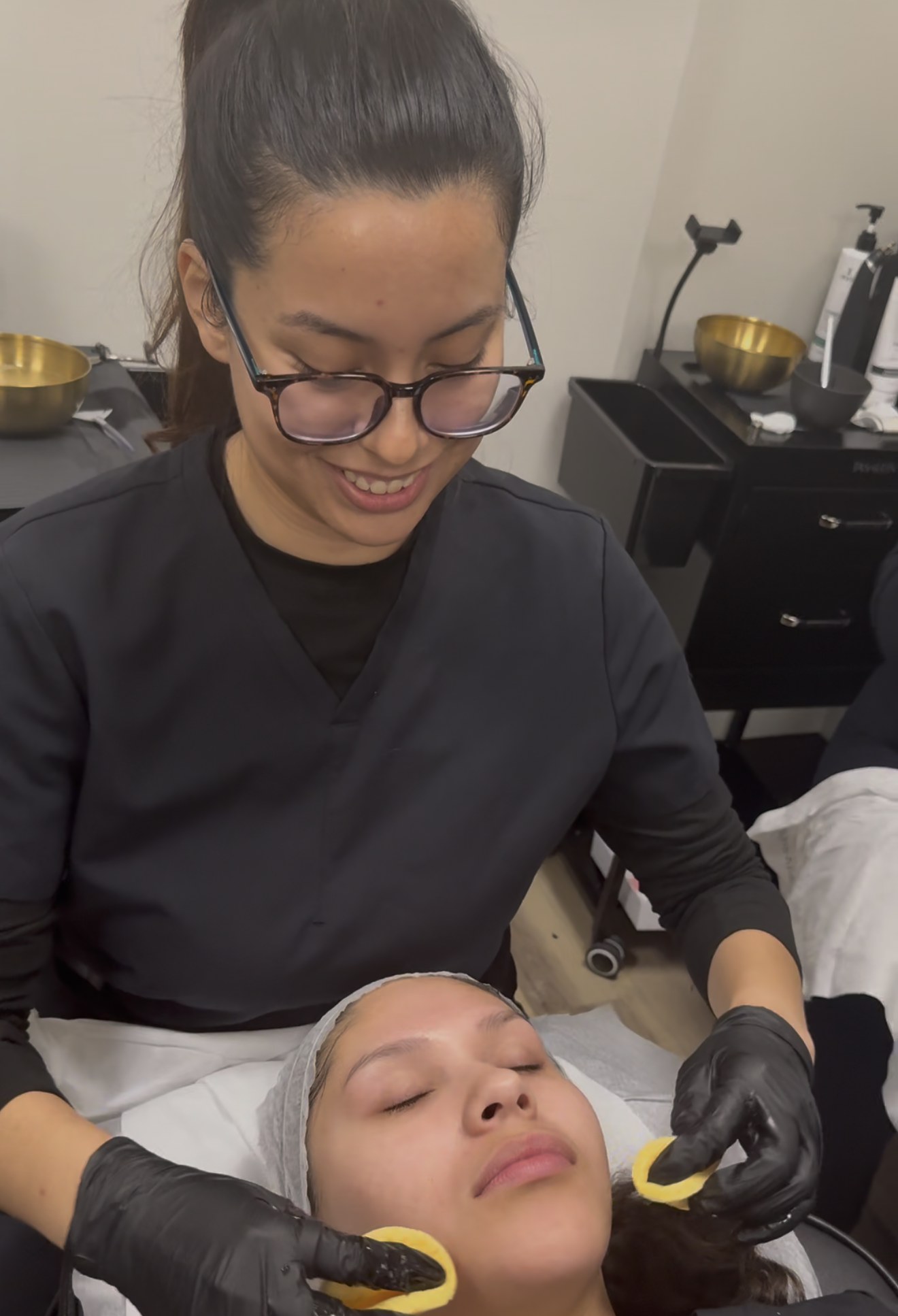 A woman with glasses and black gloves giving a facial treatment to a woman lying down with closed eyes in a spa or clinic setting.