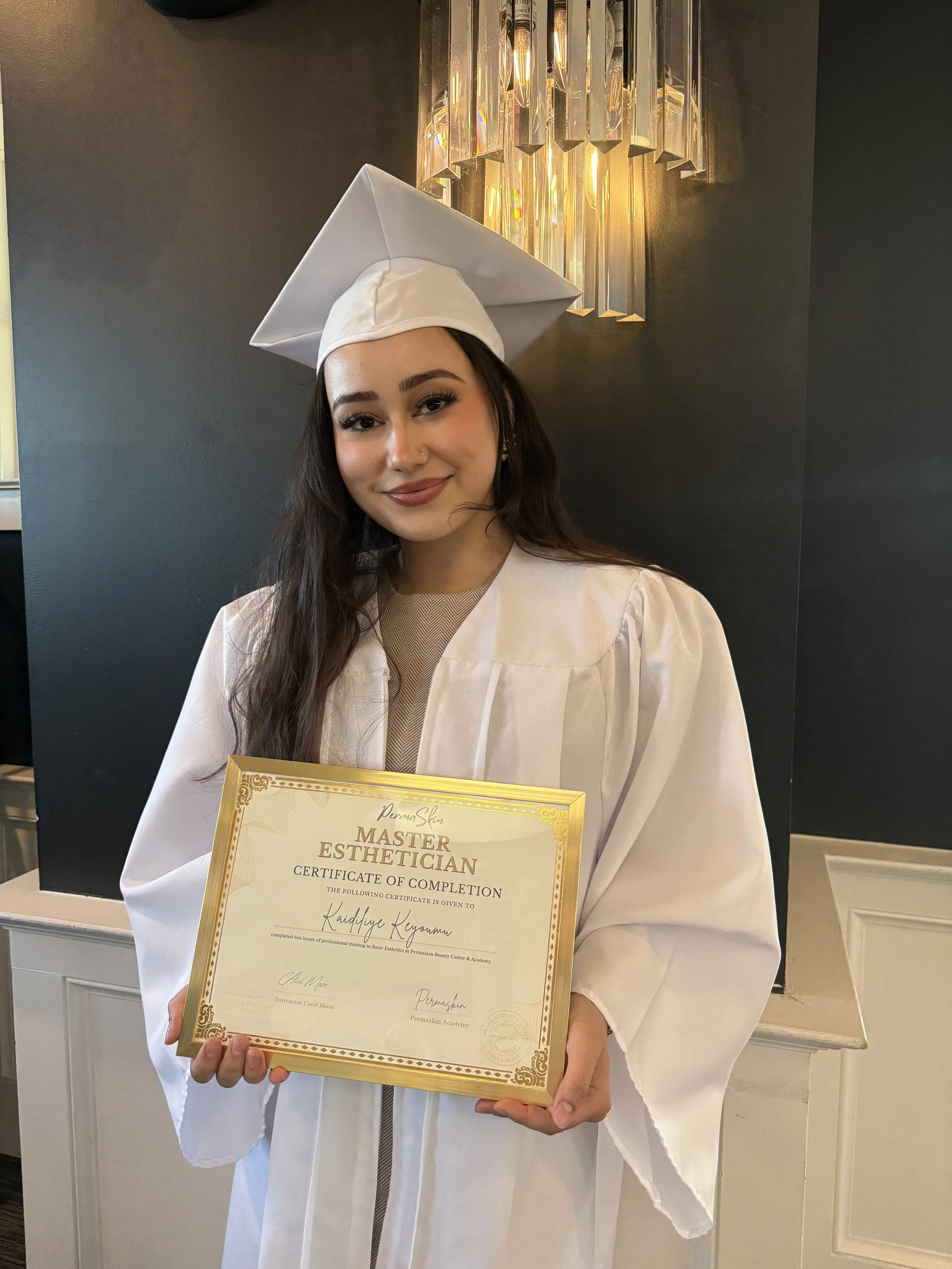 A young woman in a white graduation gown and cap holding a certificate of completion for Master Esthetician. She is smiling and standing indoors with a dark wall and modern chandelier in the background.