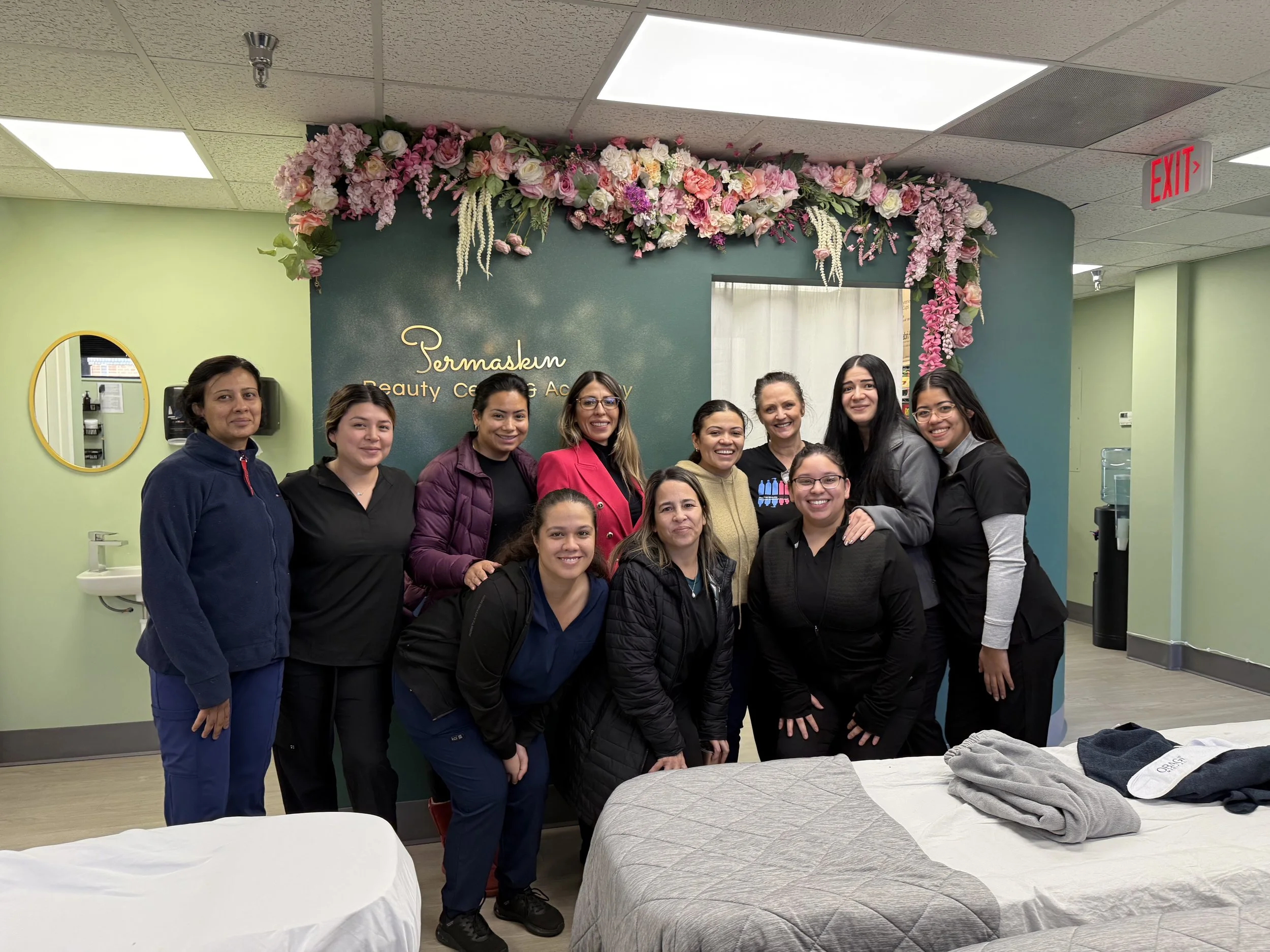 Group of ten women posing for a photo inside a salon or beauty center, with a sign that reads "Permaskin Beauty Center & Academy" on the green wall decorated with pink and purple flowers.