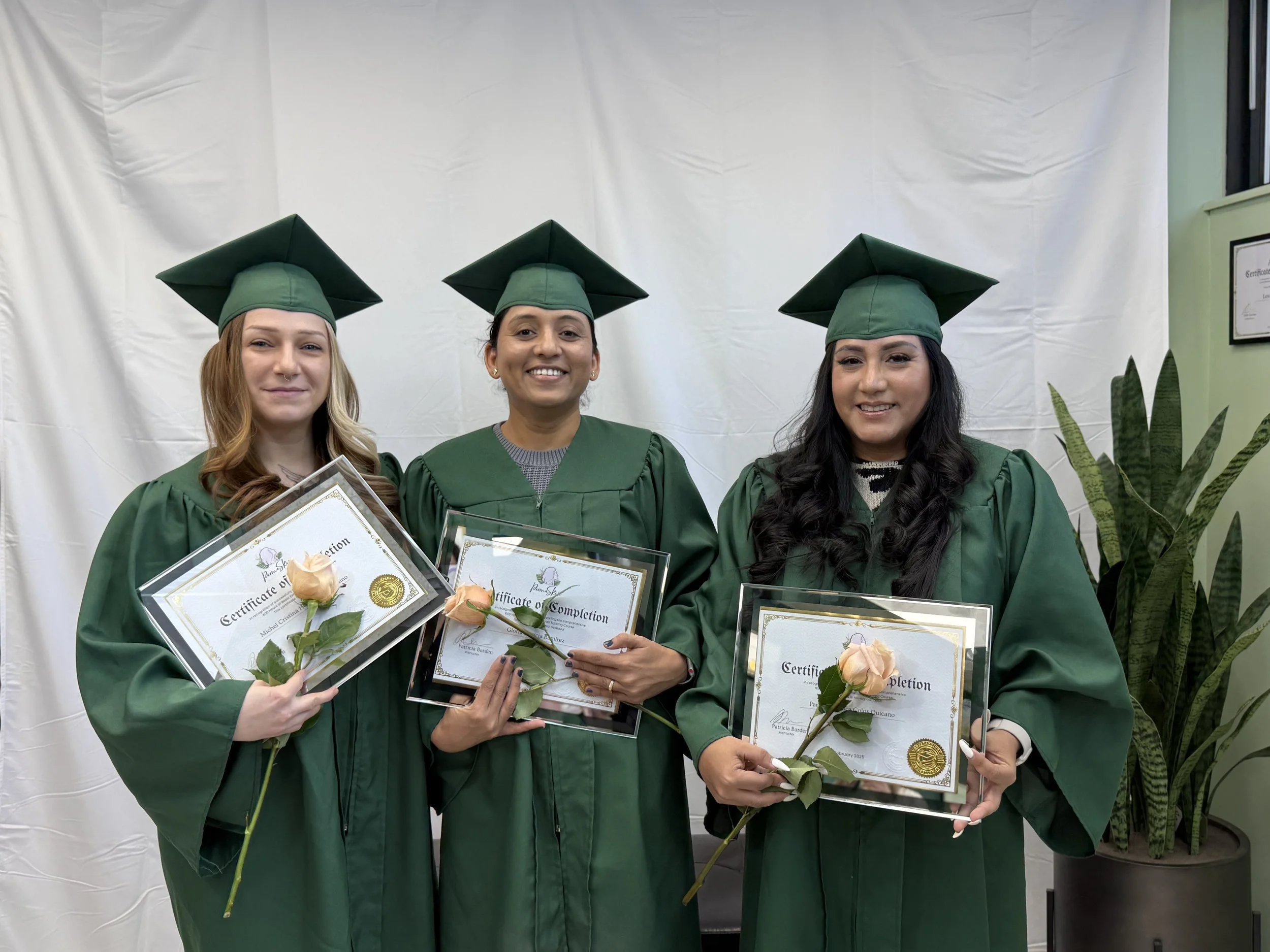 Three women in green graduation gowns and caps holding diploma certificates and pink roses, standing in front of a white backdrop and next to a potted plant.