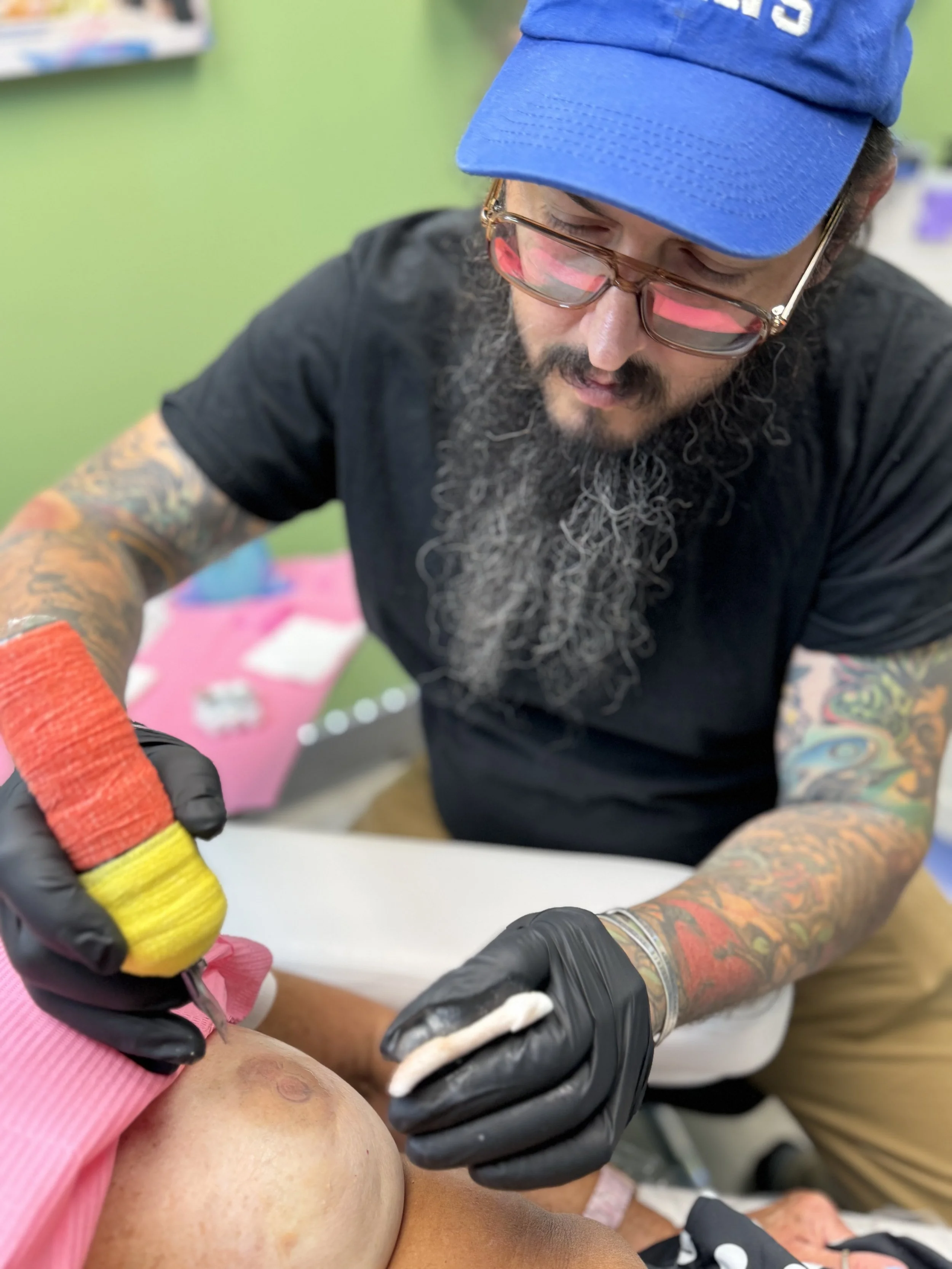 A tattooed man with glasses and a blue cap draws on a person's skin with a tattoo needle.