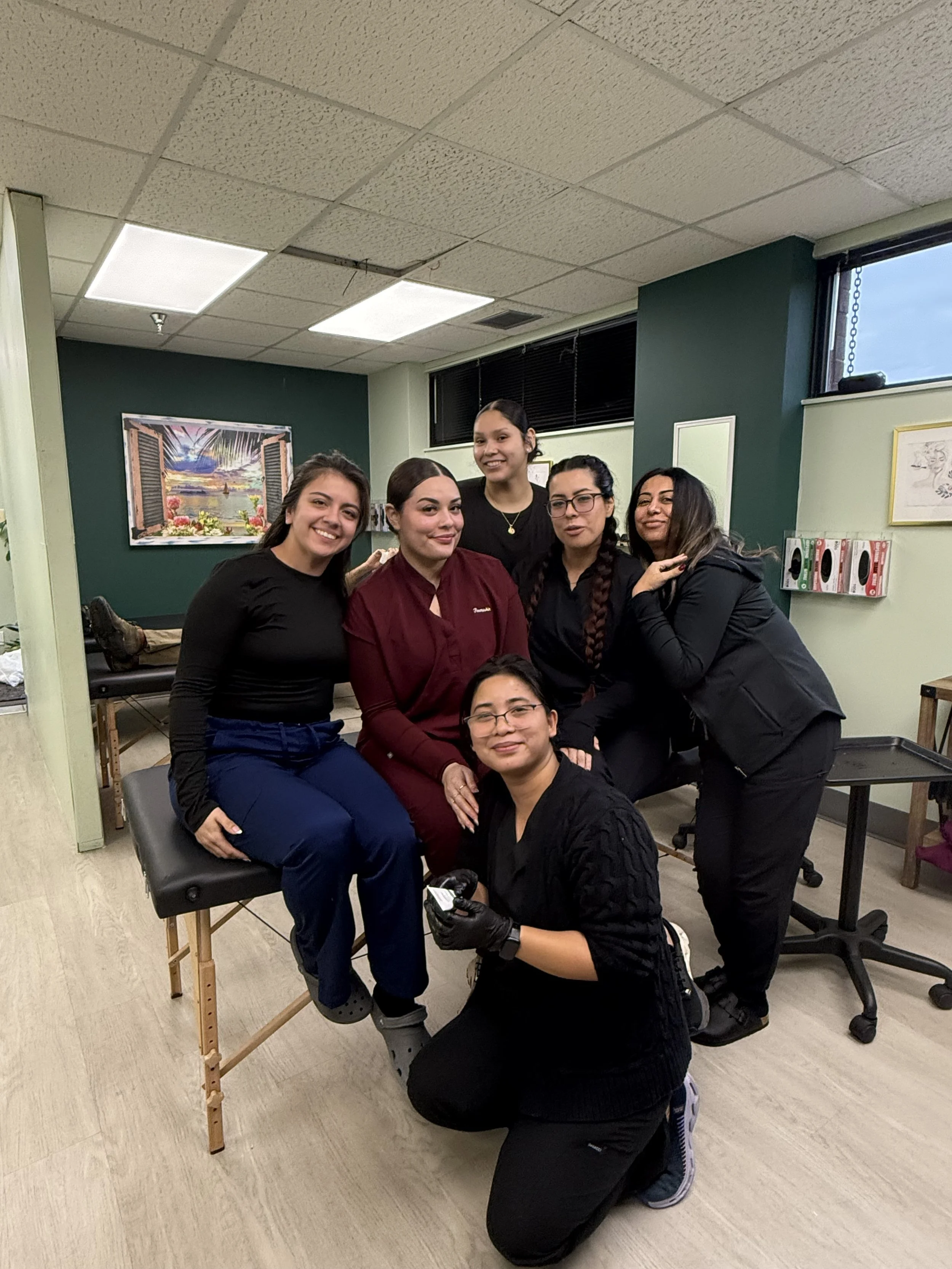 Group of six women indoors, some sitting on a massage table, others standing behind, all smiling and looking at the camera.