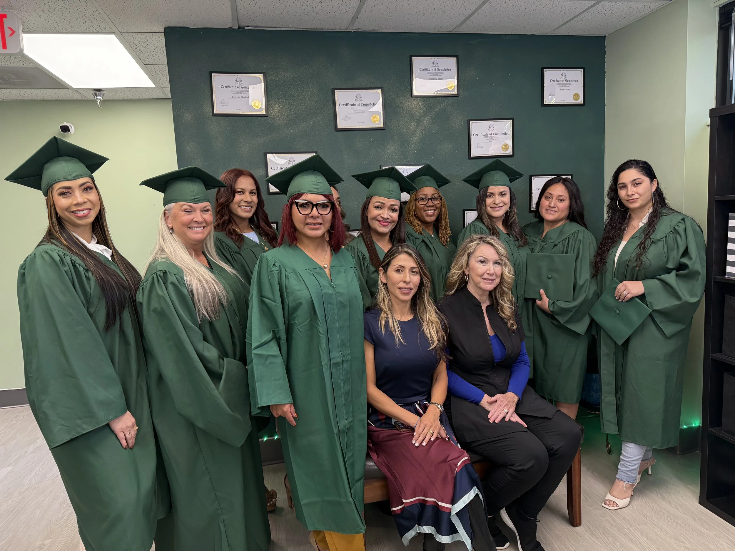 Group of women in green graduation gowns and caps, some seated and some standing, in an indoor room with certificates displayed on the wall behind them.