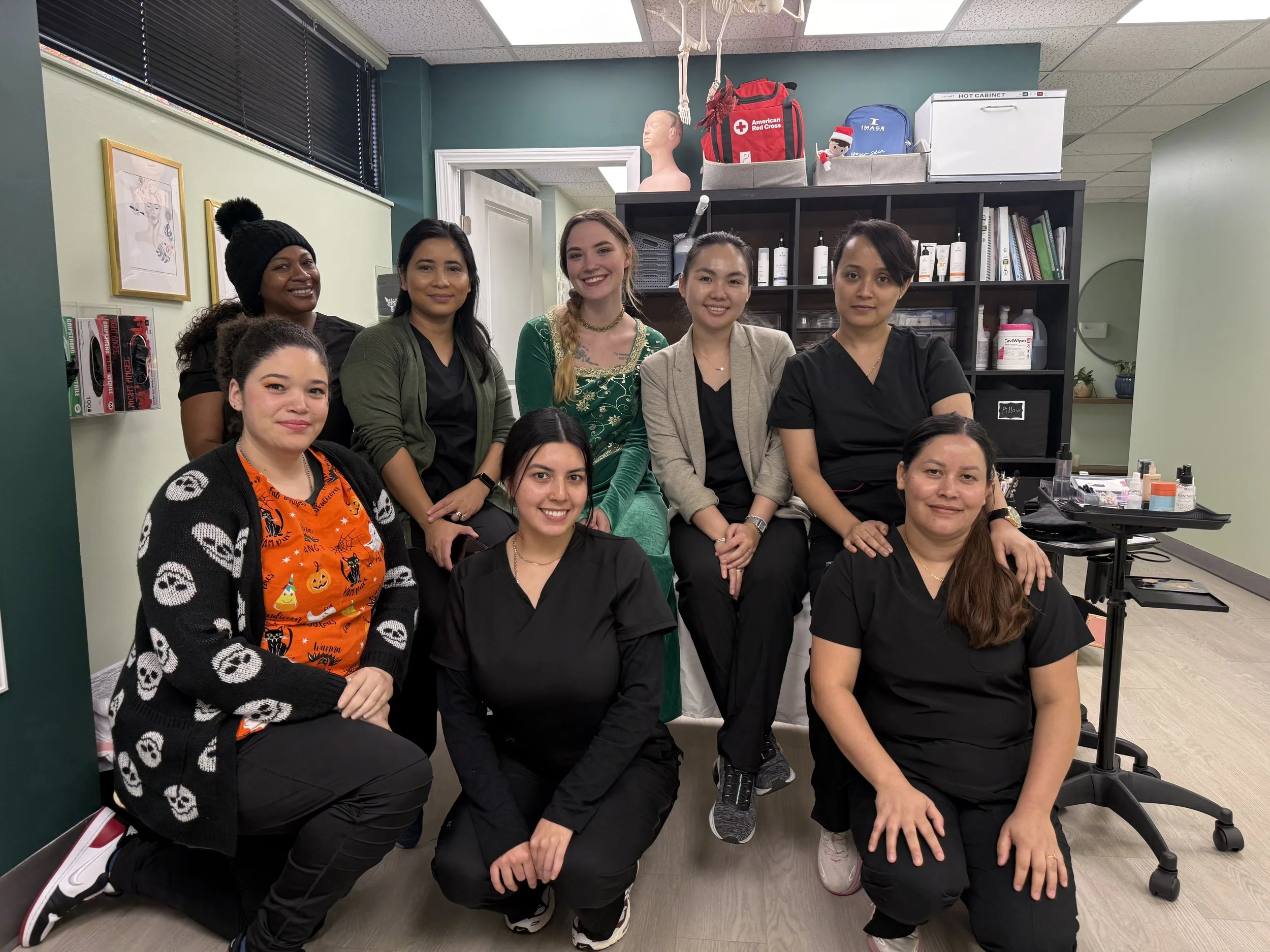 A group of nine women in medical scrubs and casual clothing posing together in a clinic or office with medical supplies and books on shelves behind them.
