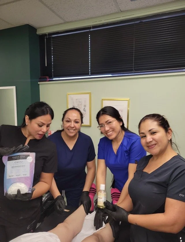 Four women healthcare workers in scrubs and black gloves smiling around a patient's leg during a medical procedure in a clinic.