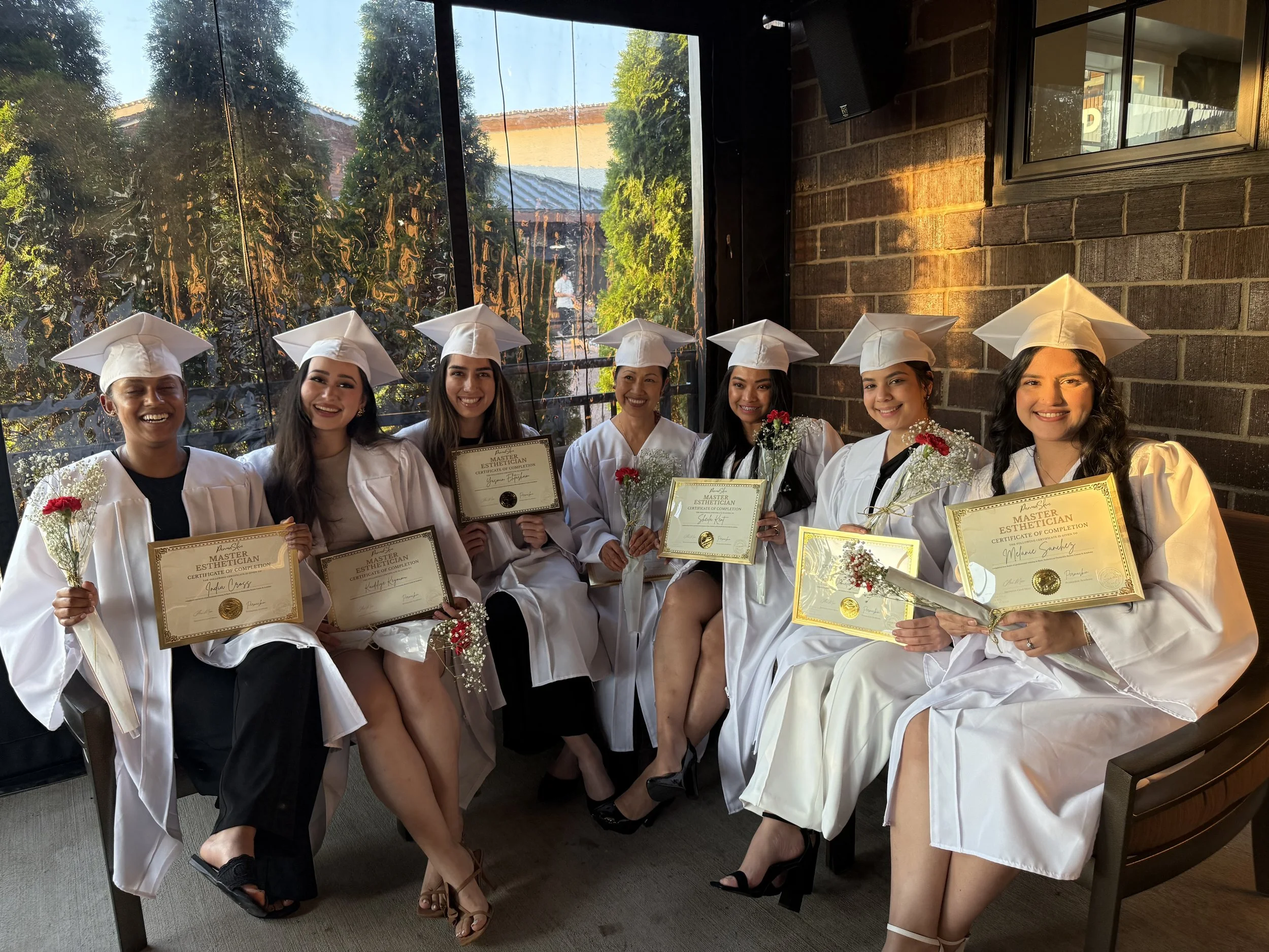 Group of seven young women dressed in white graduation caps and gowns, sitting indoors and holding certificates and flowers, celebrating graduation.
