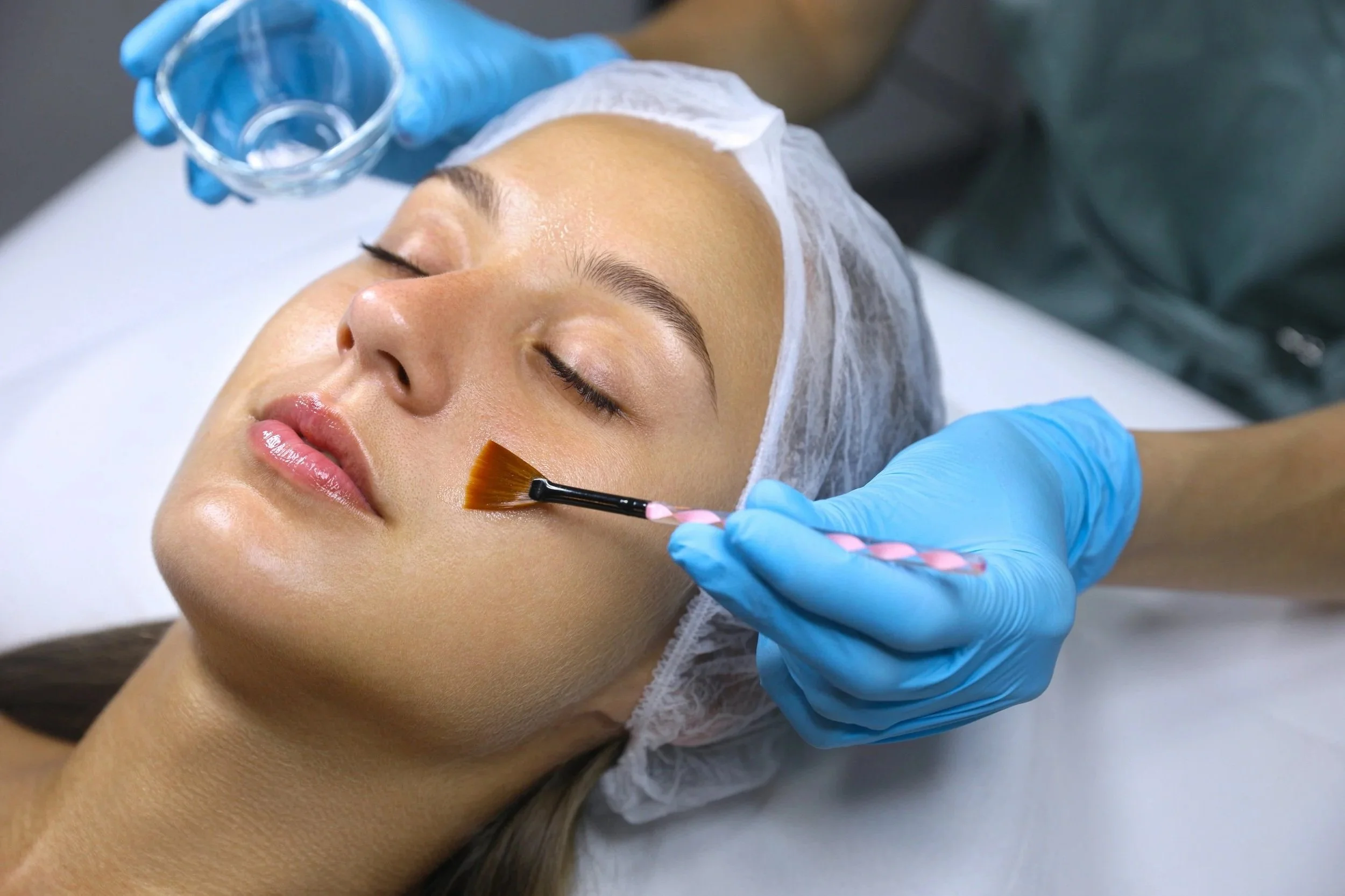 A woman receiving a facial treatment, lying down with eyes closed, while a professional applies a facial mask with a brush.