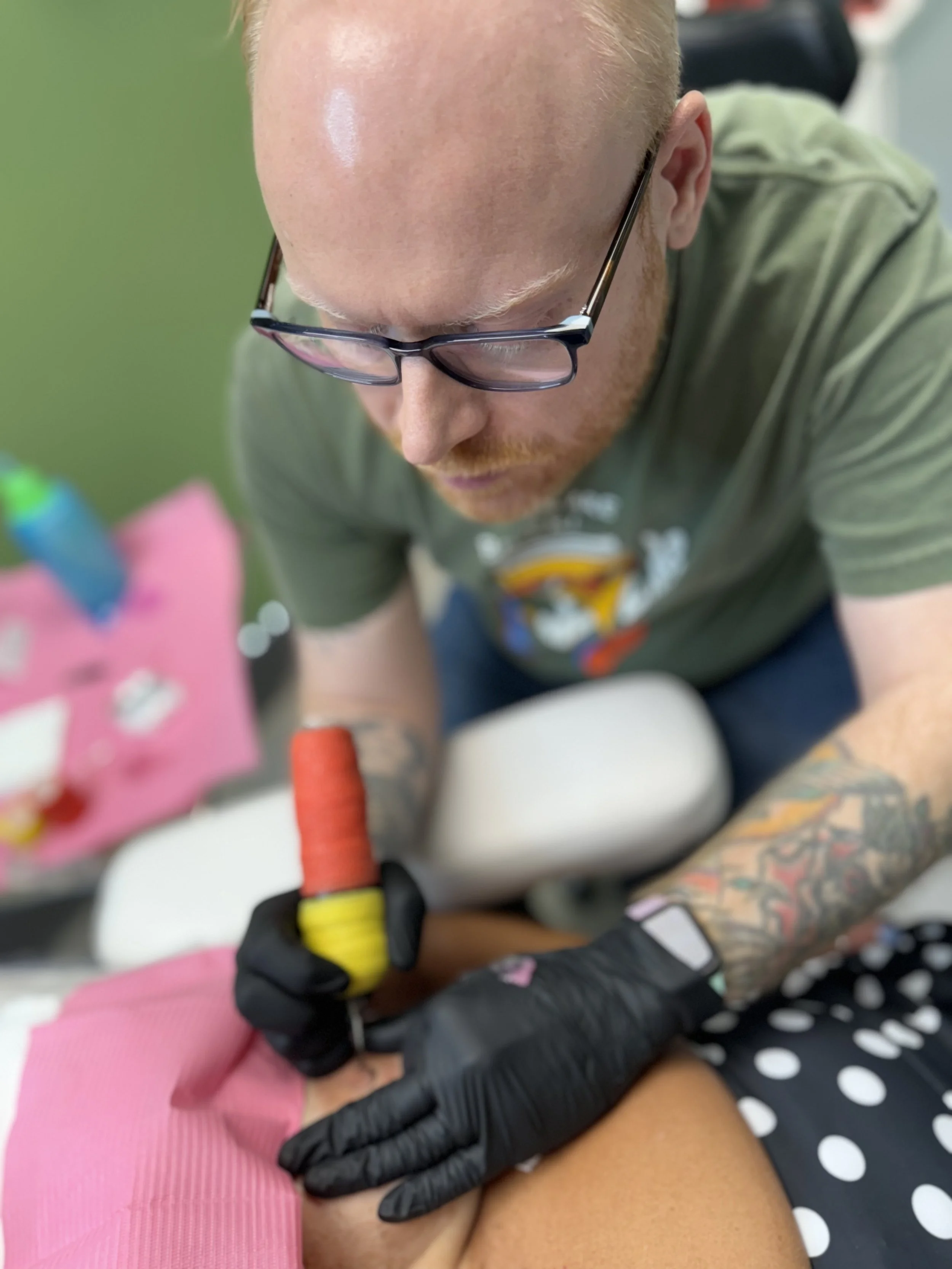 A tattooed man wearing glasses and black gloves tattooing a person's thigh with a tattoo machine.