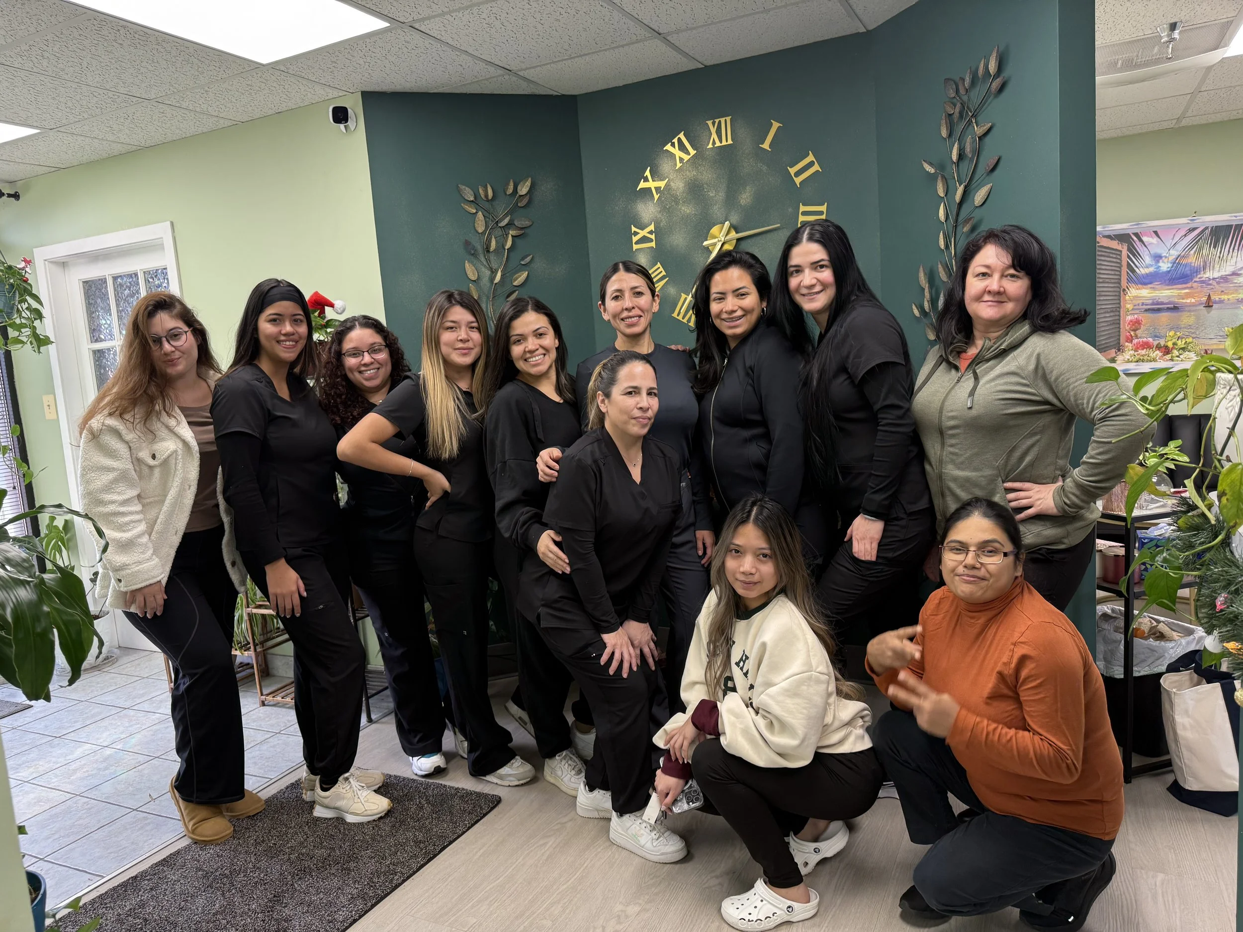 Group of 12 women posing together indoors near a green wall with a large round clock showing 2:10, decorative metal leaves, and a landscape picture. The women are smiling and some are making peace signs.
