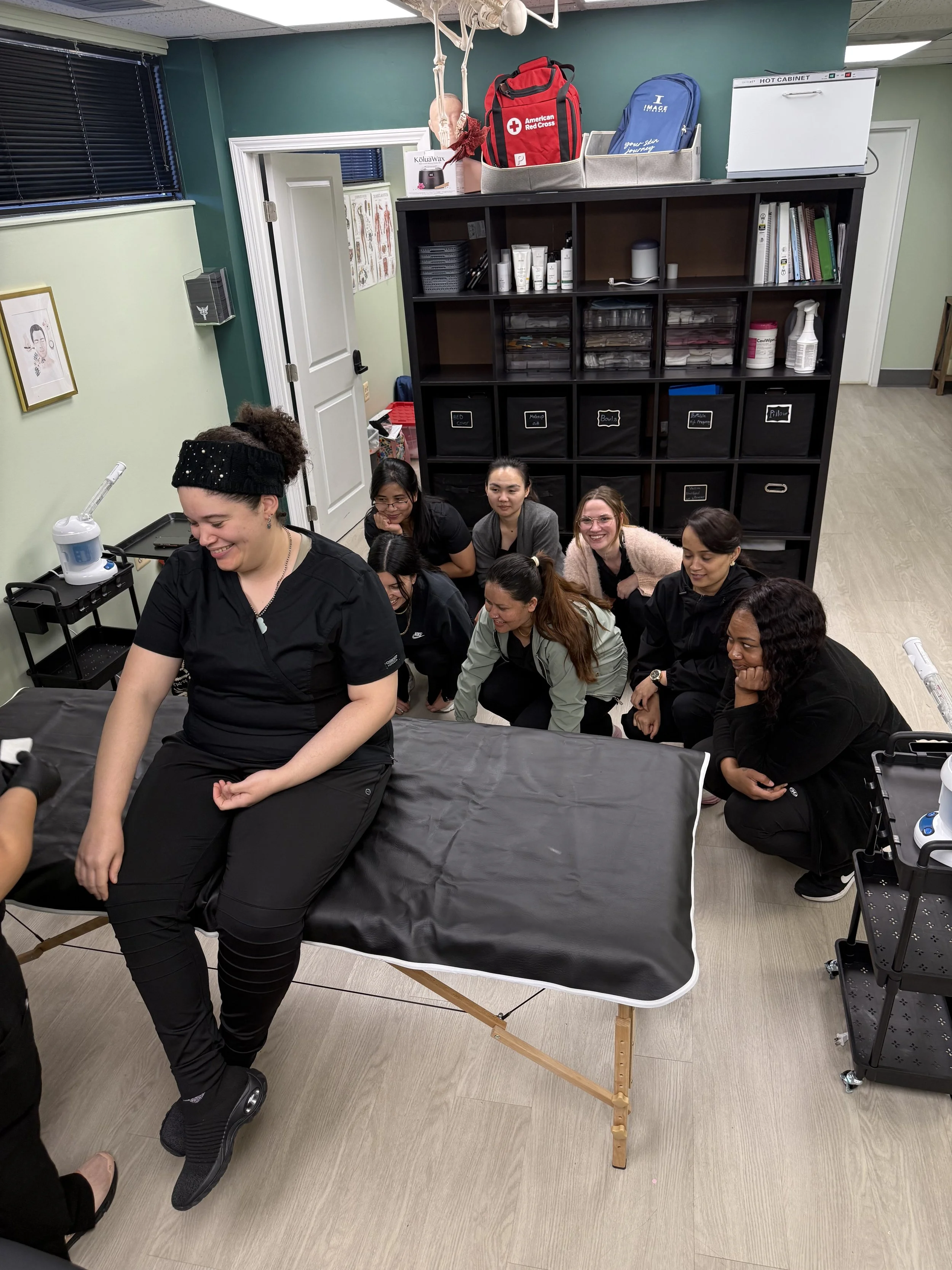 A woman in black scrubs sitting on a massage table, smiling, while a group of women watches her in a classroom or training room setting.