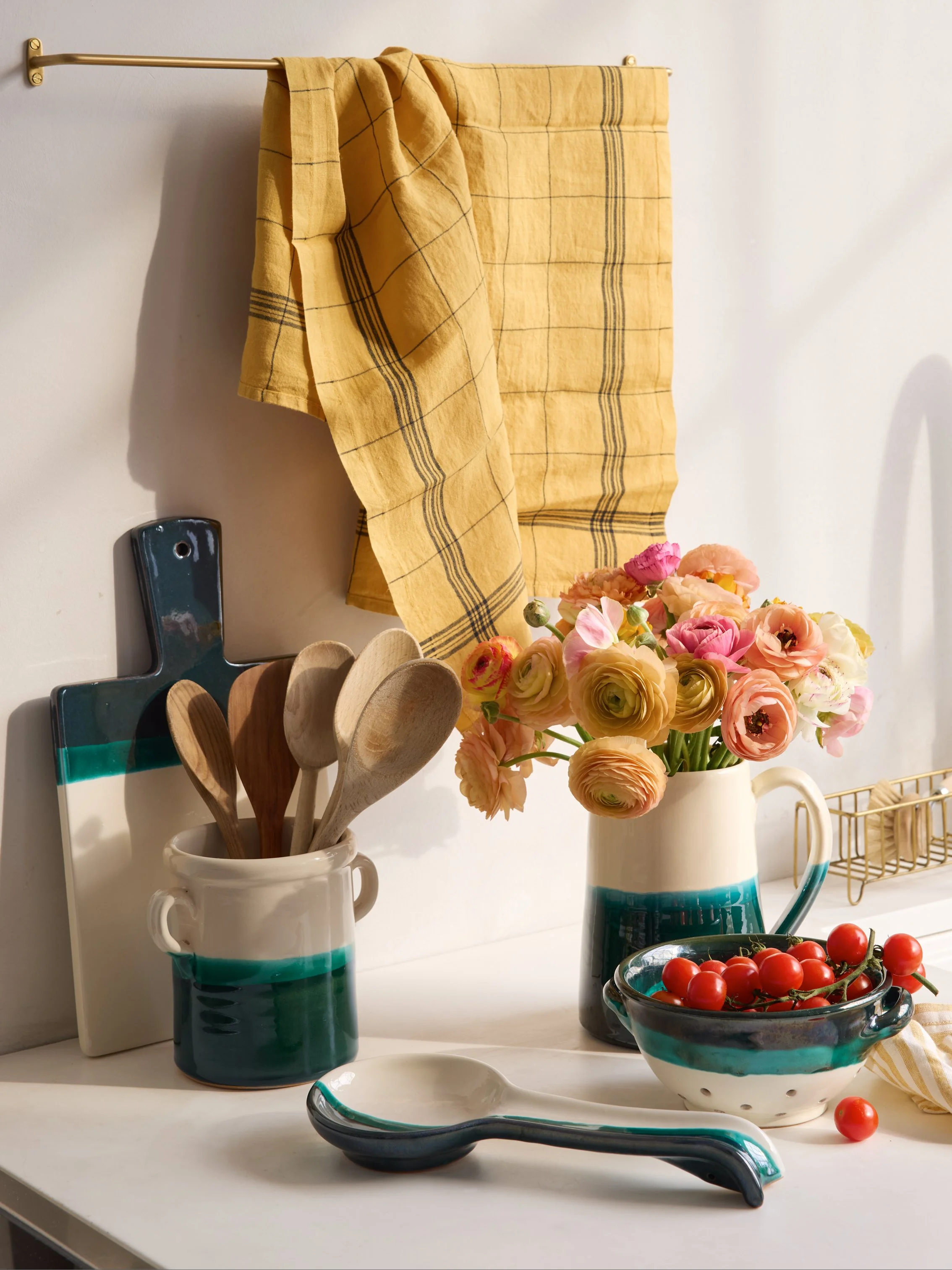 Kitchen countertop with a bowl of cherry tomatoes, a vase of pink and yellow flowers, a ceramic container with wooden spoons, a decorative spoon rest, a yellow and blue checkered dish towel hanging on a wall, and a wire dish rack.