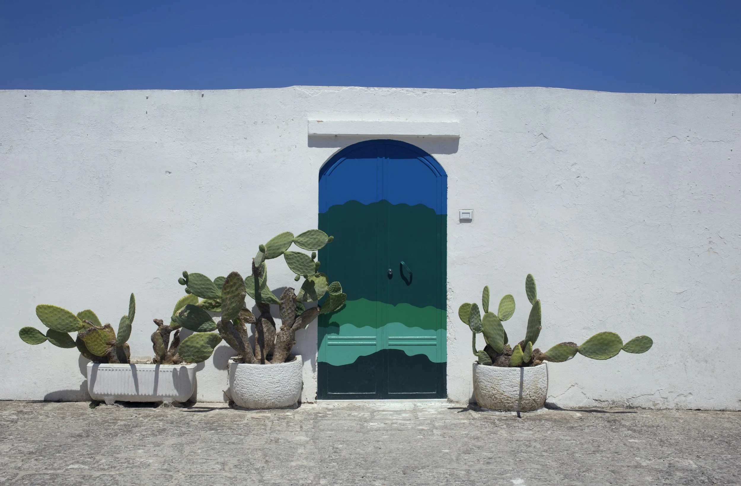 White stucco wall with colorful painted blue and green door, flanked by three potted cactus plants on a concrete ground under a clear blue sky.