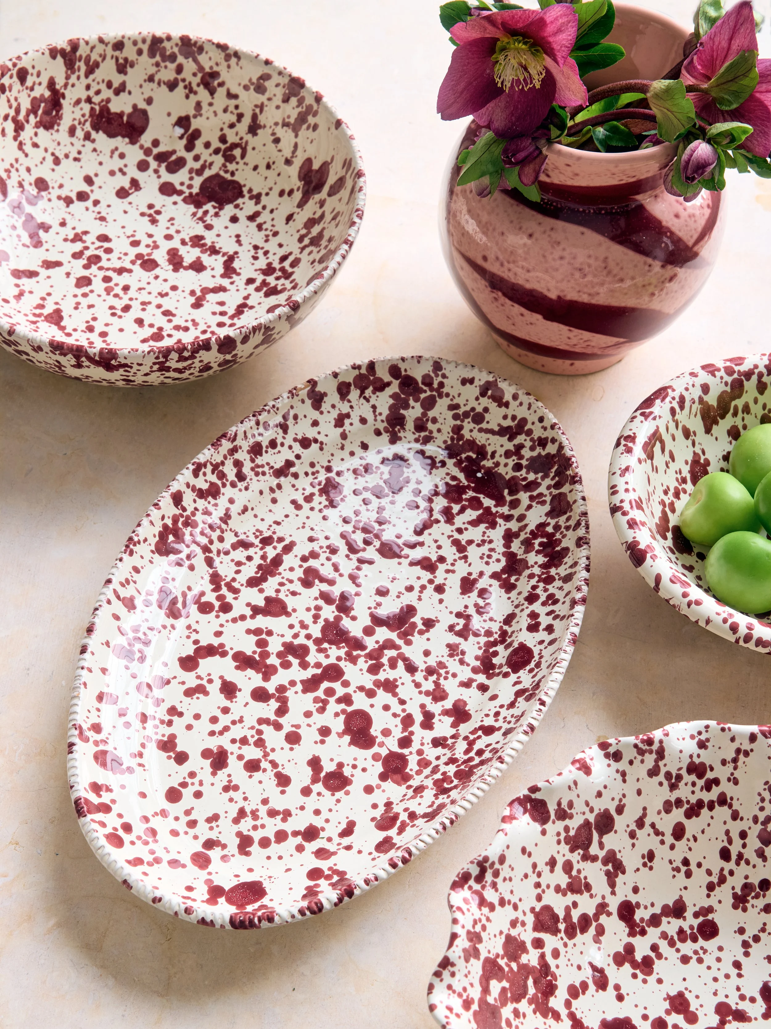 Ceramic bowls with red and white splatter patterns, a pink and red striped vase with pink flowers, and a small bowl of green grapes on a light-colored surface.