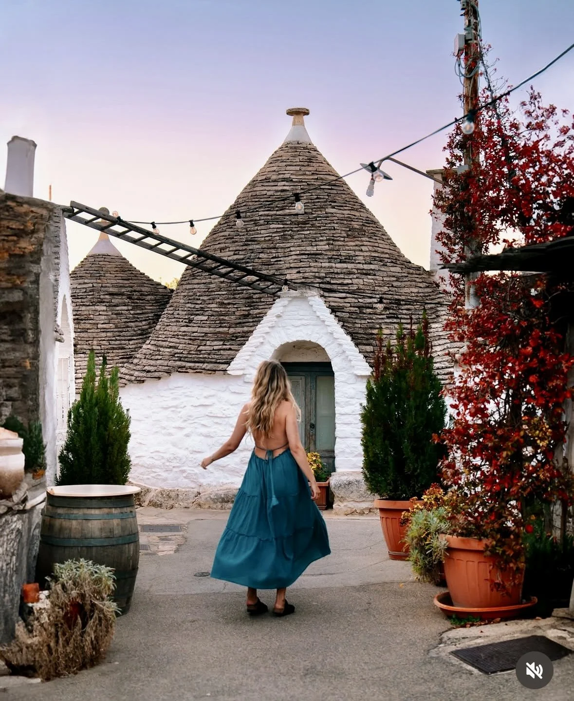 A woman in a teal dress walking past Trulli-style white stone houses with conical roofs, surrounded by potted plants and autumn foliage.