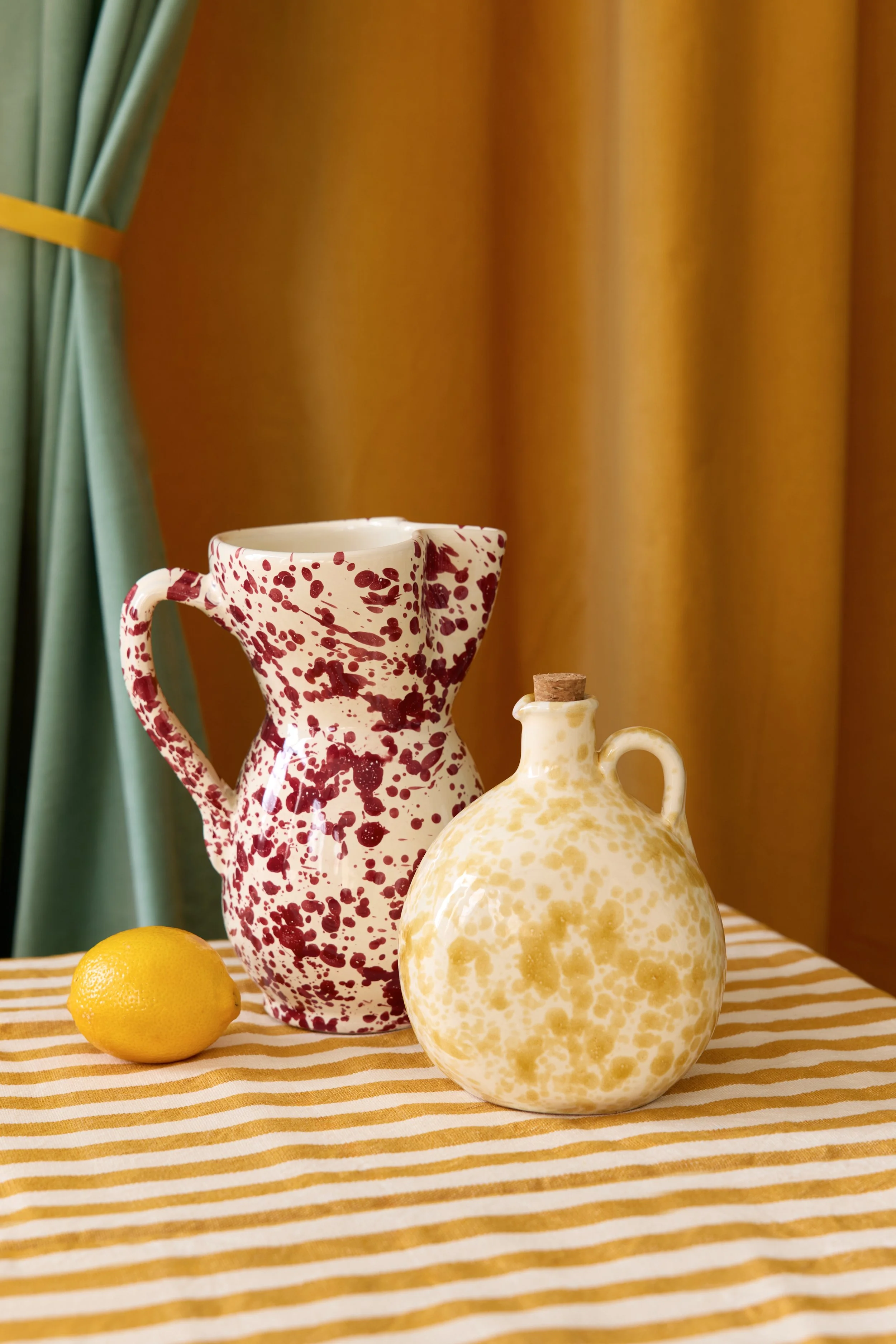 A table with a yellow and white striped tablecloth, displaying two ceramic vases and a lemon. The first vase is tall with a handle, decorated with red and white splatter pattern. The second vase is smaller, round with a handle, featuring a yellow and white speckled pattern and a cork stopper. A lemon is placed on the table near the vases. In the background, there is a yellow curtain and part of a green curtain.