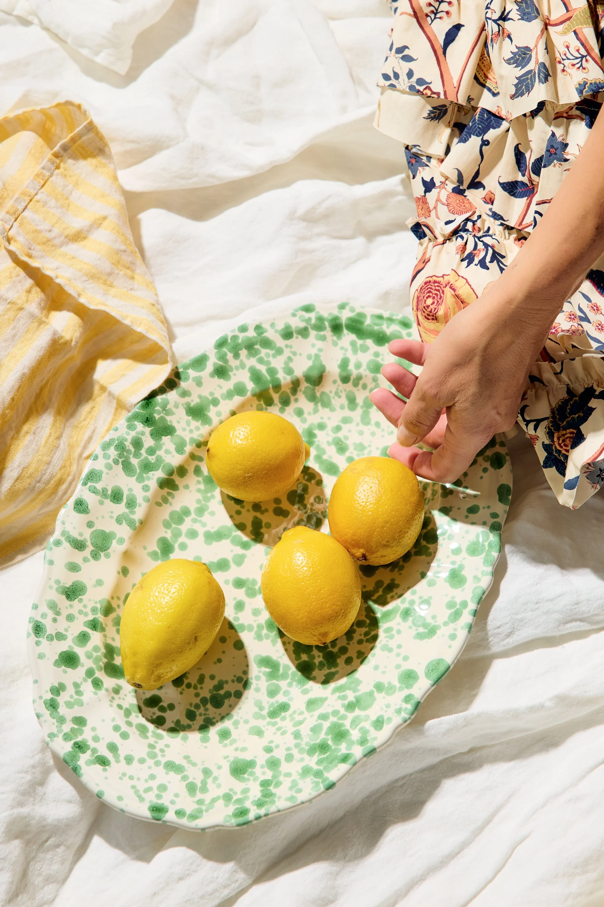 Four lemons on a green and white speckled ceramic plate, with a person's hand reaching for one, placed on a white textured surface with cloths nearby.