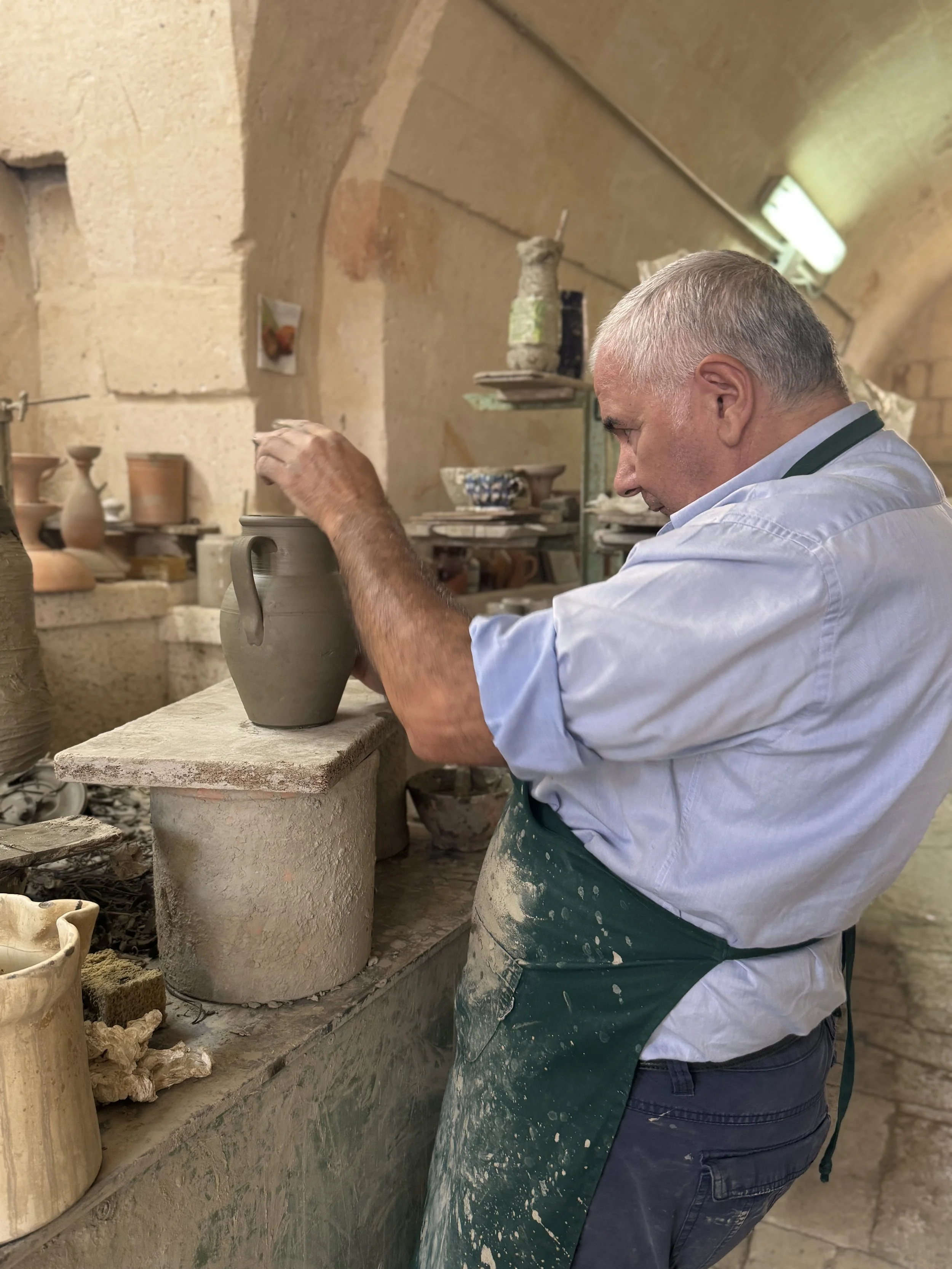 A man with short gray hair working on a pottery wheel in a pottery studio, wearing a white shirt and a green apron covered in clay.