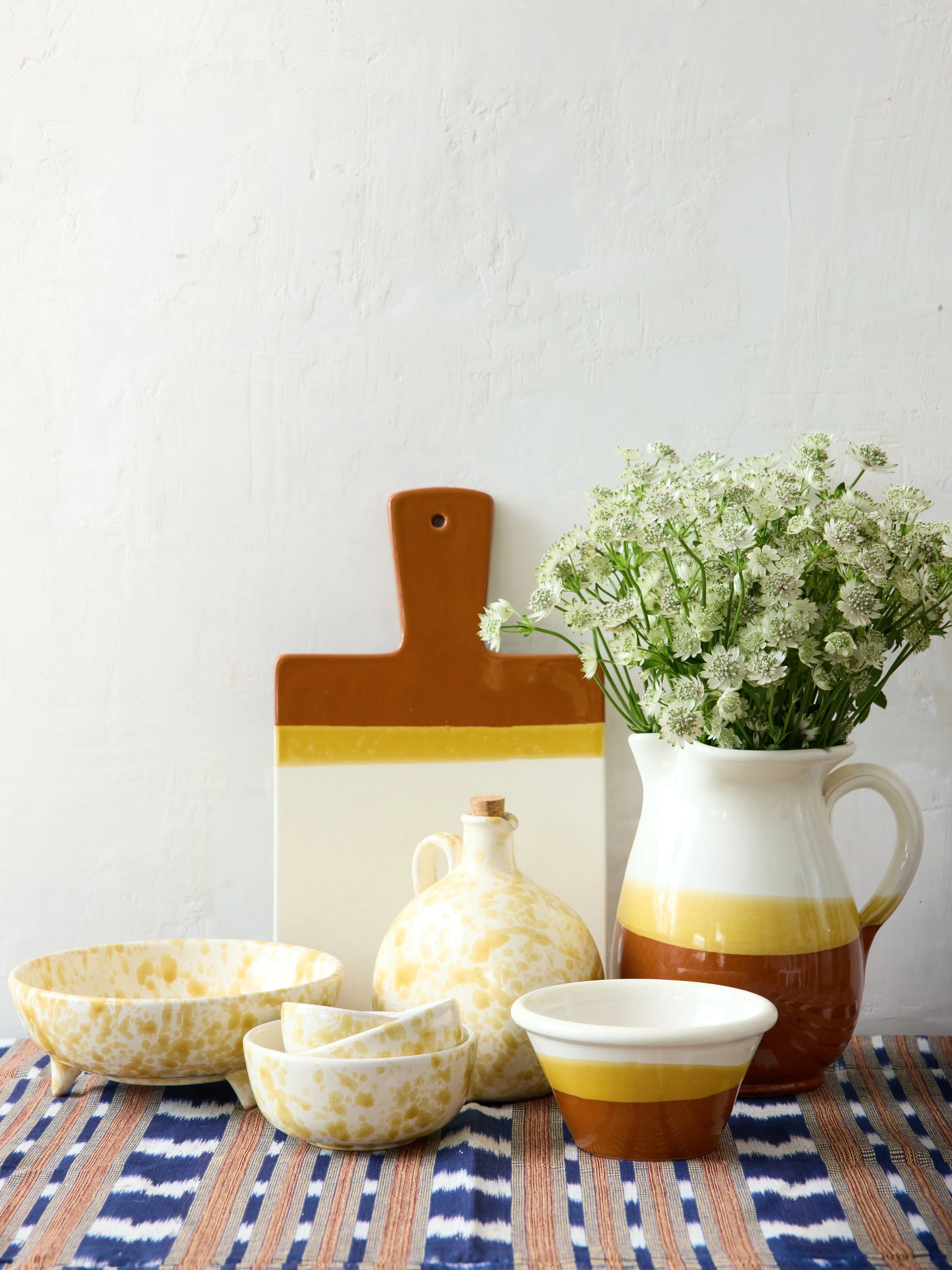 Ceramic kitchenware set including a bowl, two smaller bowls, a small jug, and a large pitcher with a handle, all featuring white, yellow, and brown colors, placed on a woven striped tablecloth. A large white ceramic vase holds a bouquet of white and 