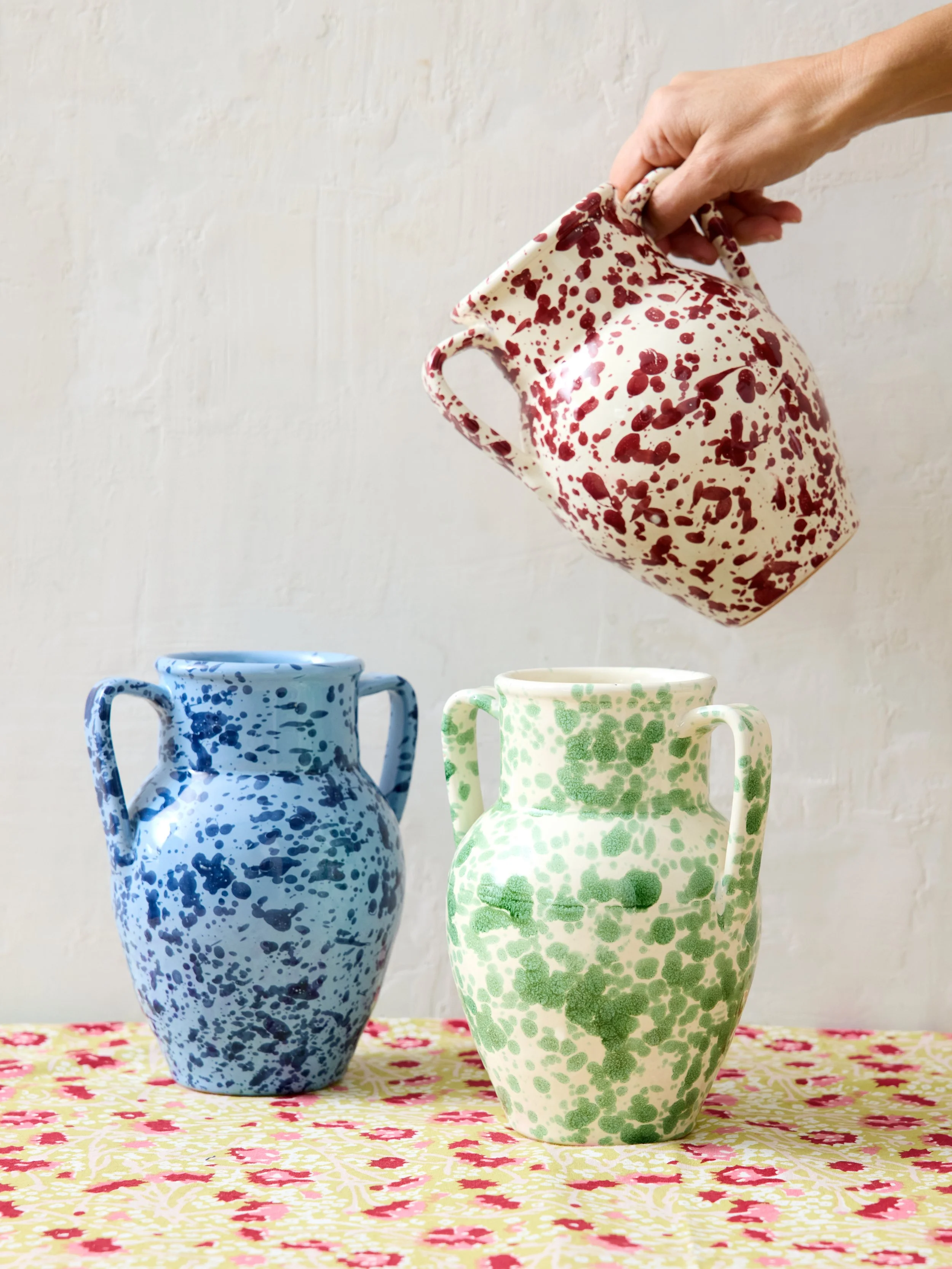 Hand holding a speckled red and white ceramic pitcher above two speckled ceramic vases, one blue and one green, on a floral tablecloth with a plain white wall background.