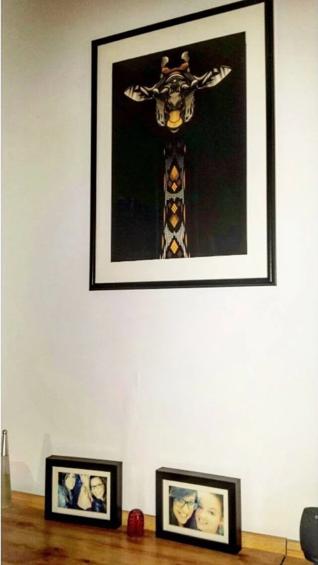Framed tribal art print of a giraffe with geometric patterns hangs on a white wall. Below it on a wooden table are two framed photos of smiling women and a small red decorative item.